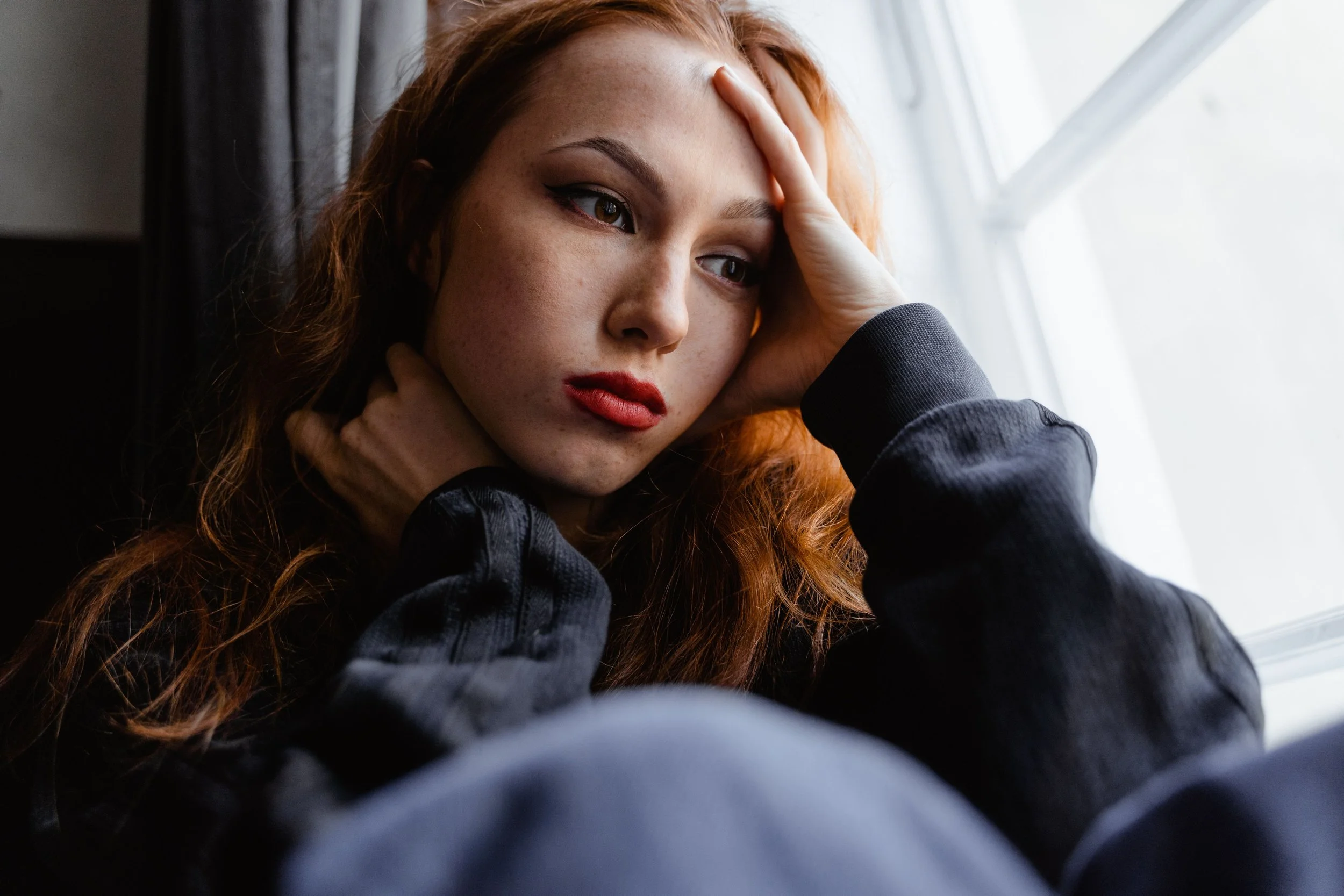 A woman with long, wavy red hair and freckles has her hand on her forehead, looking out a window with a contemplative expression.