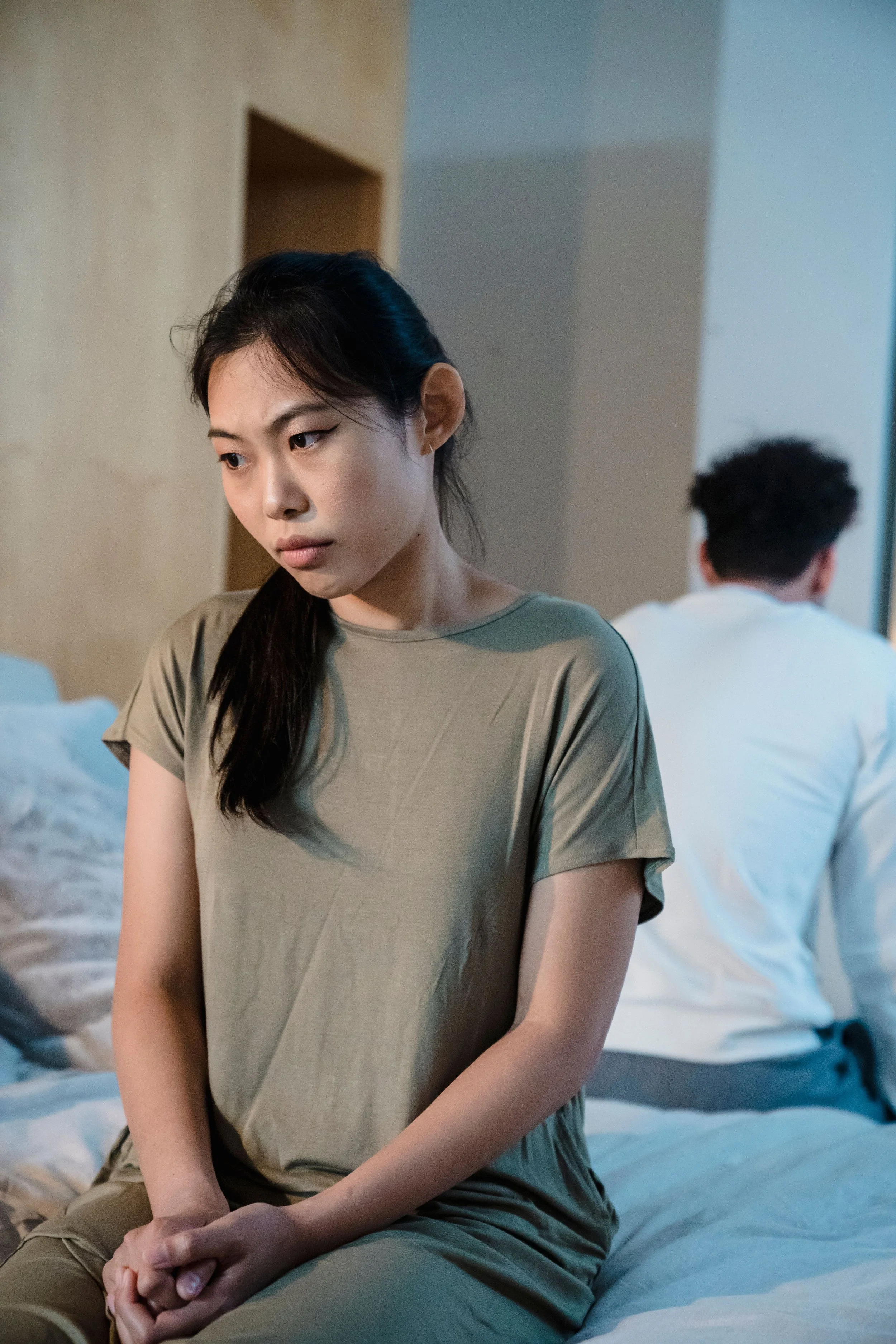A woman sitting on a bed with her hands clasped, looking down with a thoughtful expression, while a man with his back to the camera sits on the same bed facing away. The scene suggests a tense or reflective moment in a bedroom.