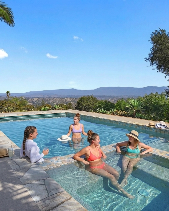 Four women are enjoying a sunny day in a swimming pool surrounded by nature and mountains in the background.