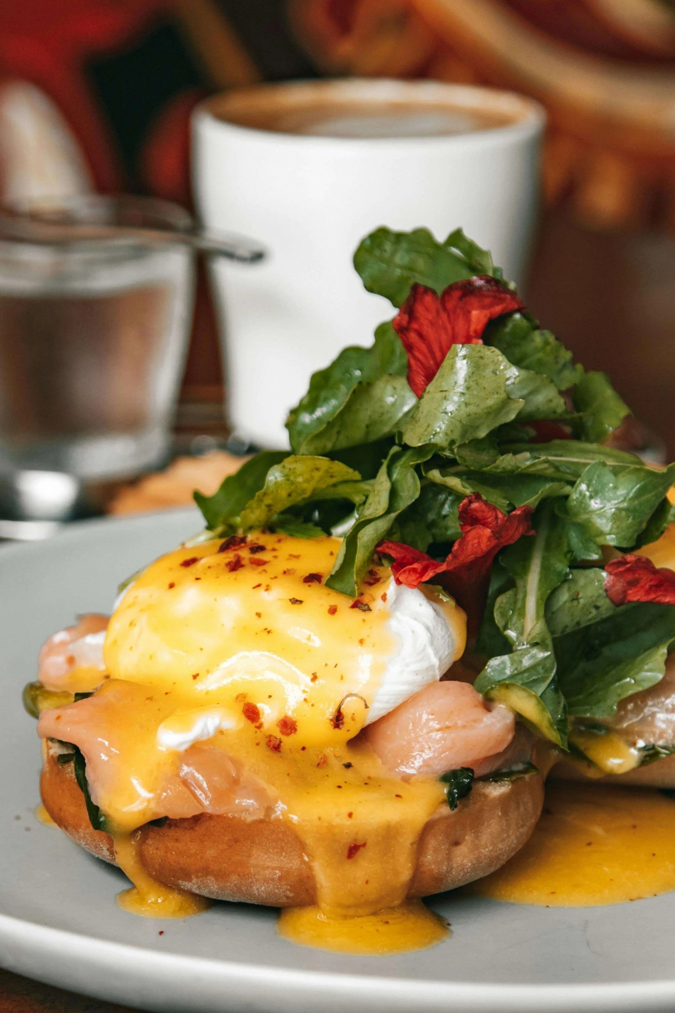 A plate of Eggs Benedict topped with hollandaise sauce, served with leafy greens and red flower petals, with coffee and a glass of water in the background.