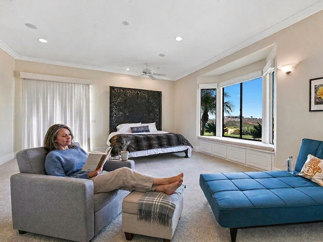 A woman relaxing on a light gray sofa, reading a book, in a spacious bedroom with a large bed, a blue lounge chair, and big windows showing a sunny outdoor scene with palm trees.