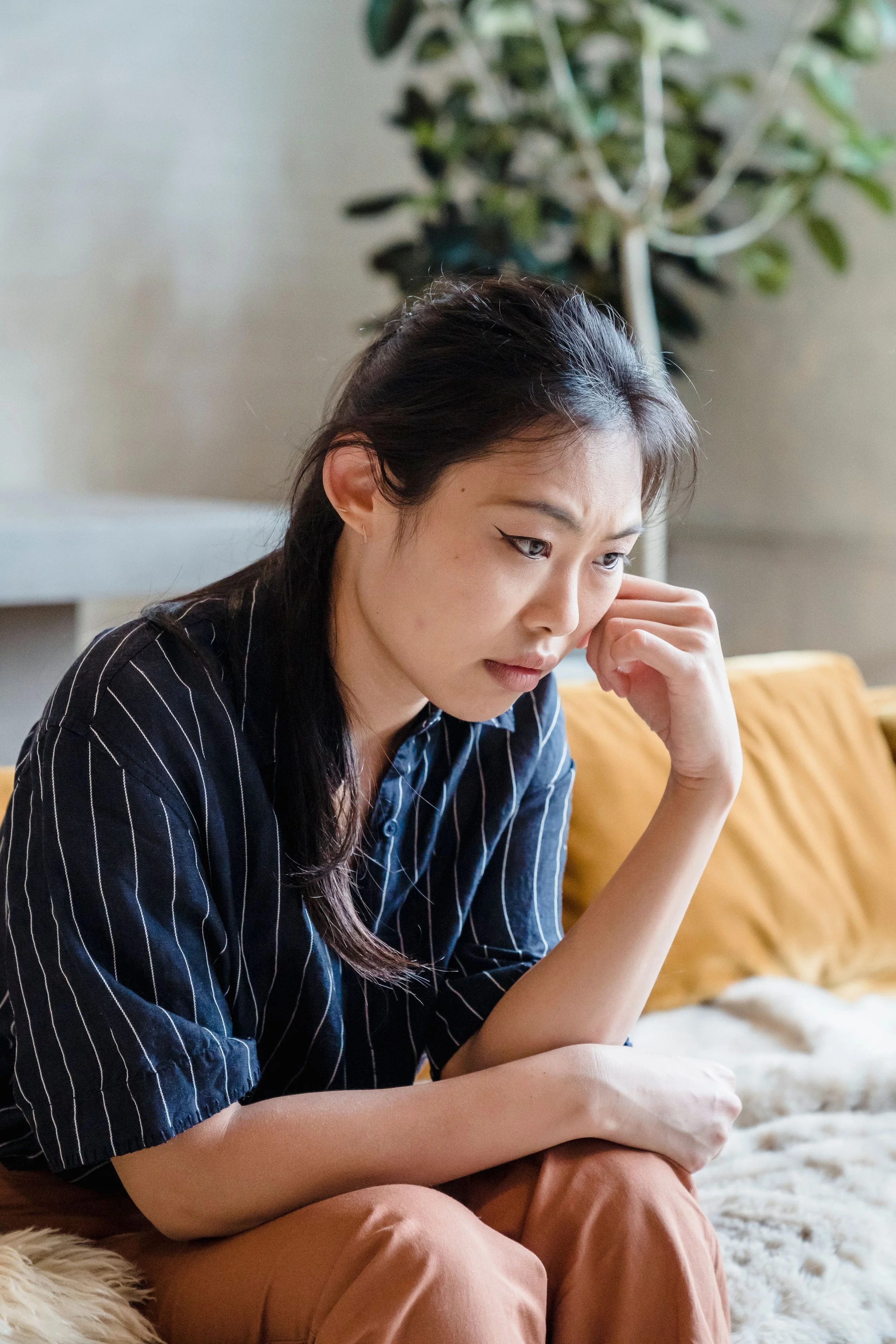Young woman sitting on a furry rug, deep in thought, with a large green houseplant in the background.