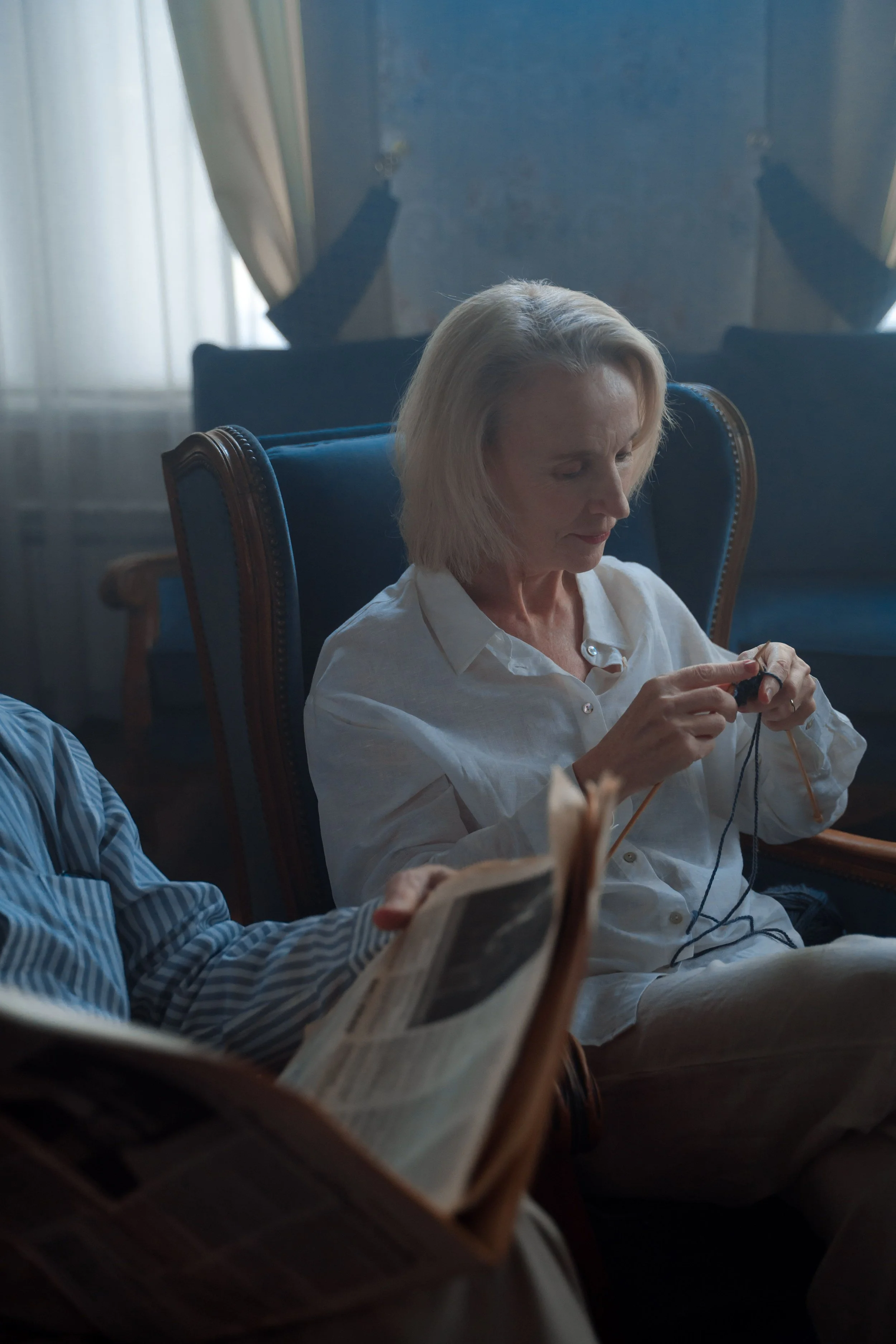 A woman with blonde hair and a white shirt seated in a blue armchair, looking down at knitting in her hands with a calm expression. Sunlight filters through the curtains in the background.