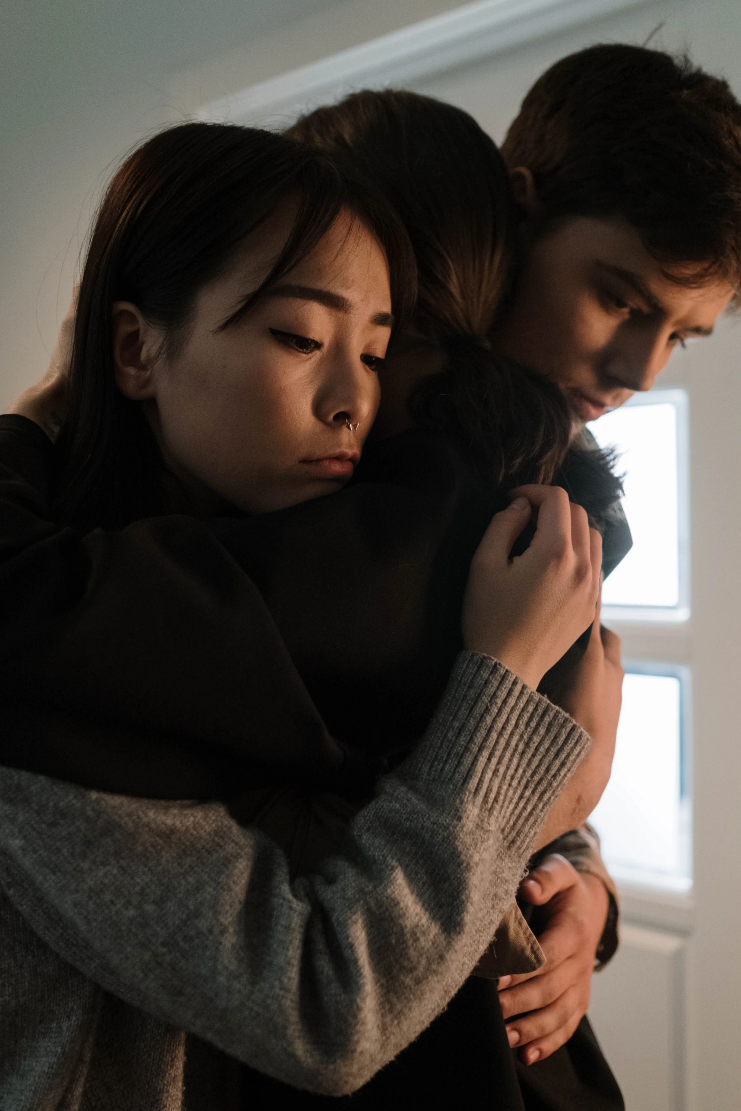Three people embracing each other, close-up of their shoulders and heads, indoors near a window.
