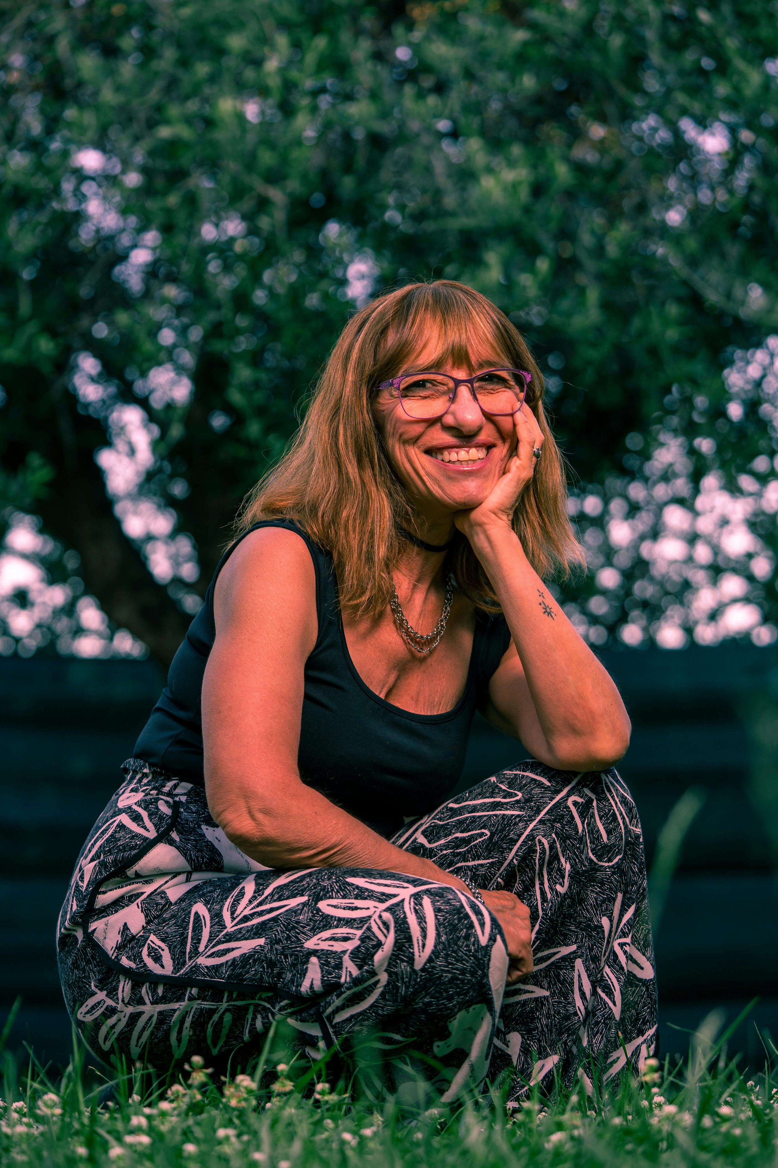 A smiling woman with glasses and shoulder-length hair, wearing a black sleeveless top, patterned pants, and jewelry, crouching outdoors on grass with trees in the background.