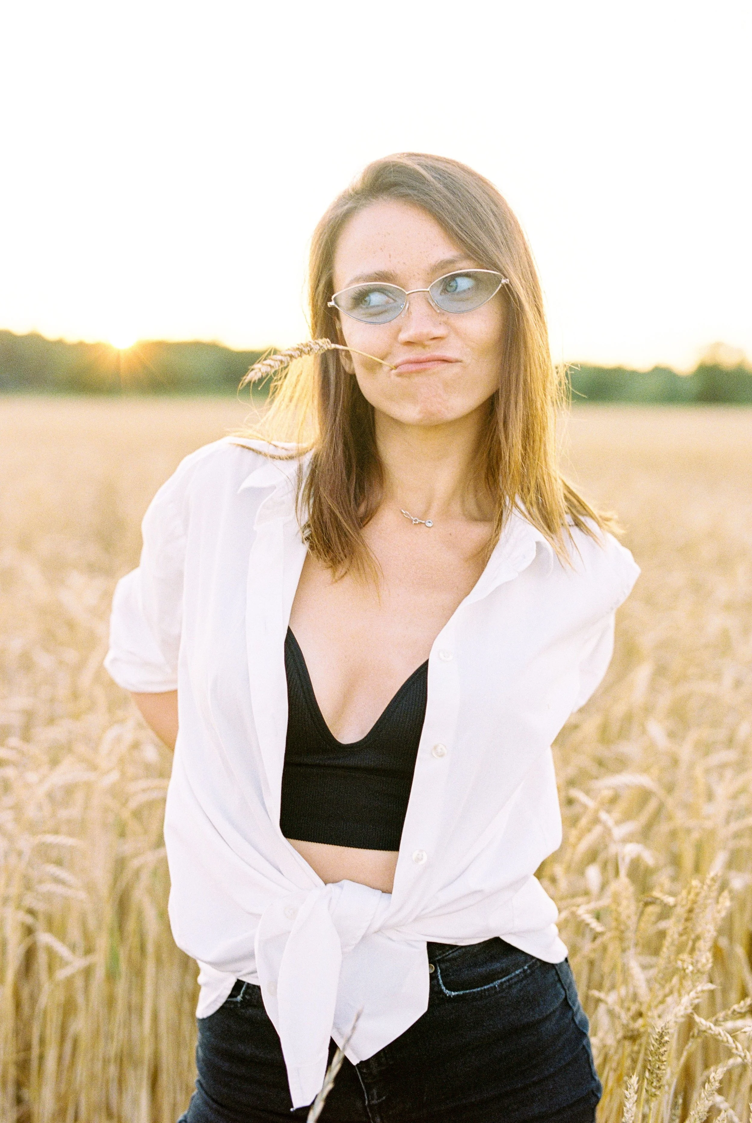 A young woman with red hair and glasses standing in a wheat field at sunset, wearing a white shirt and black crop top.