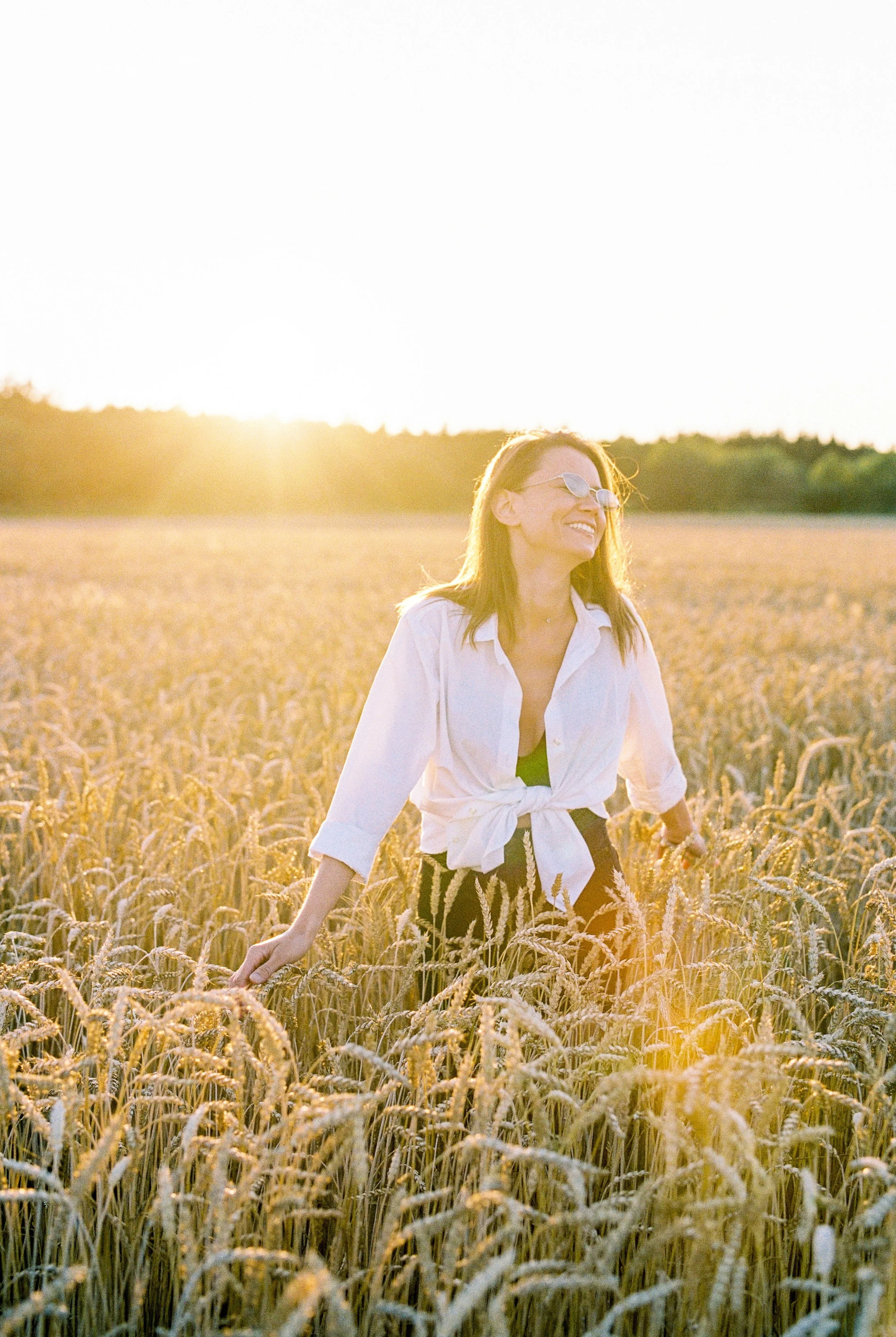 A woman smiling and walking in a wheat field during sunset, wearing sunglasses, a white shirt with rolled-up sleeves, and a black skirt with a floral pattern.