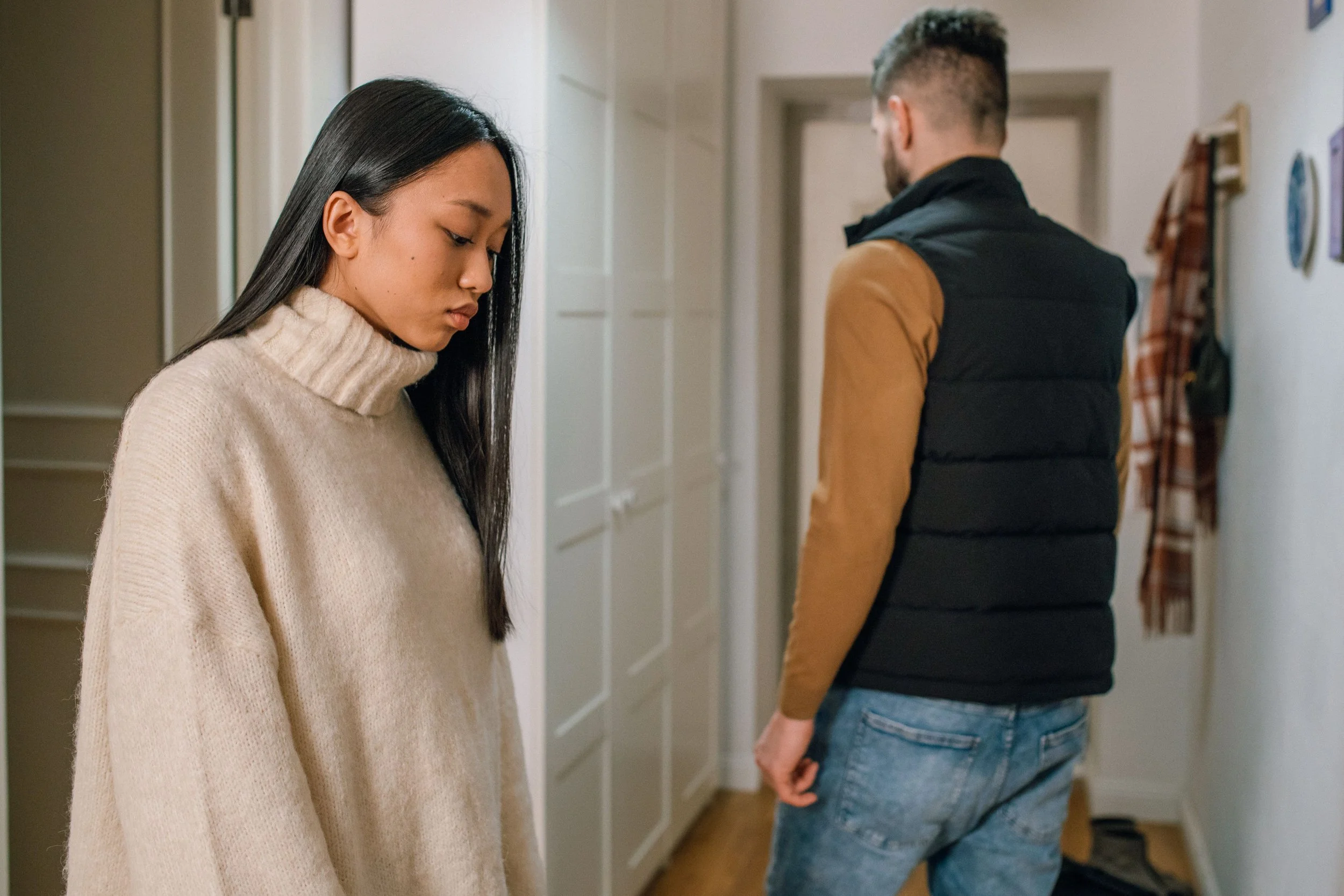 A young woman with long black hair and a beige sweater looking down, standing beside a man with short hair, beard, wearing a black vest over a brown long sleeve shirt, and blue jeans, who is facing away and leaning against a wall in a narrow hallway.