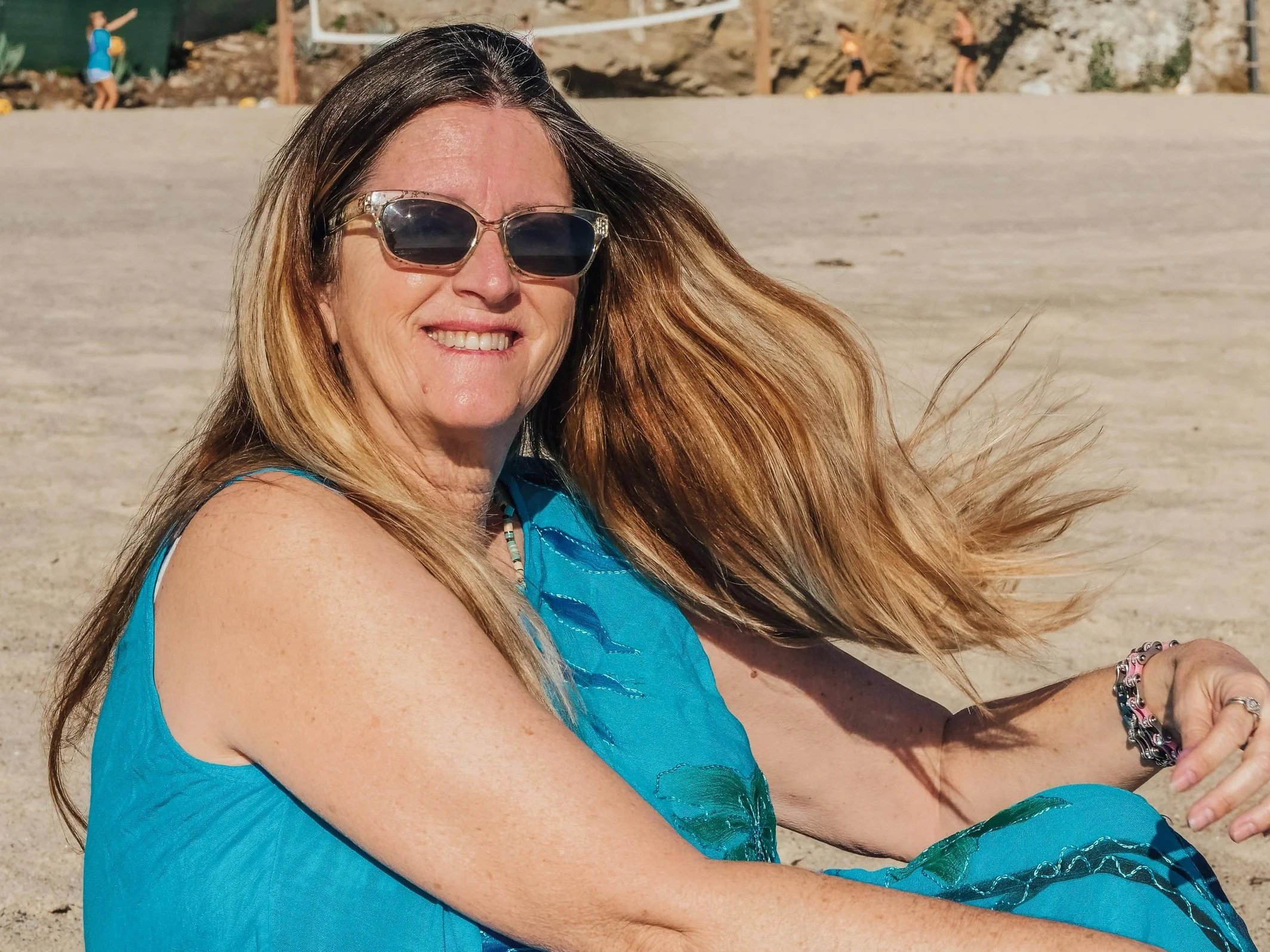A smiling woman with long brown hair wearing sunglasses and a blue sleeveless dress, sitting on a sandy beach with wind blowing her hair.