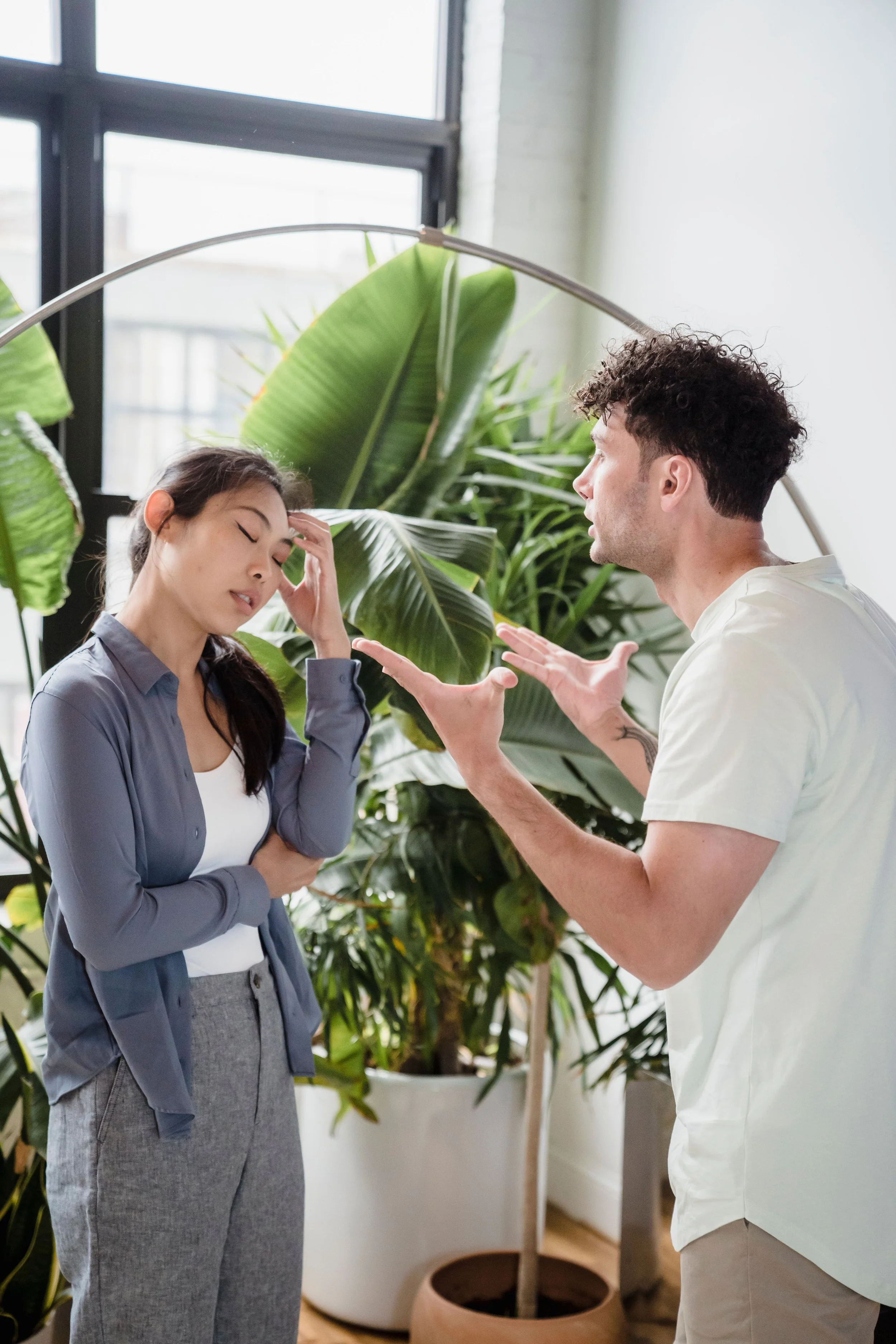 A man and a woman having an animated discussion indoors, with large green houseplants in the background.