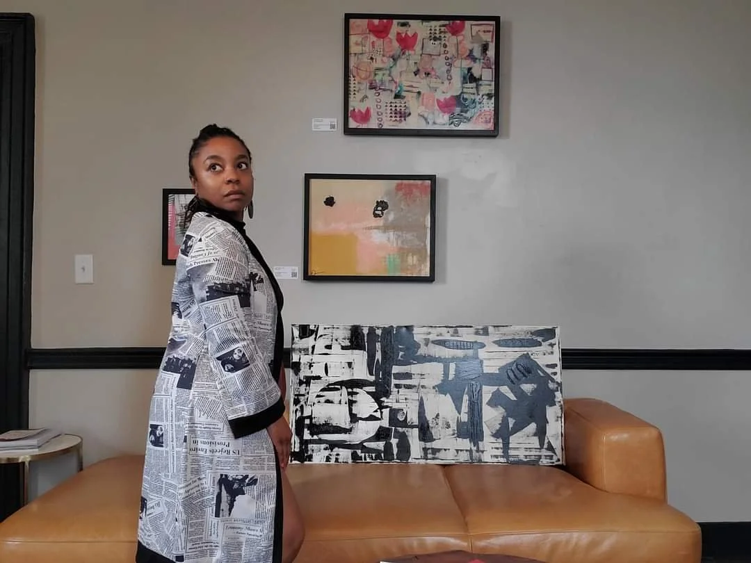 A woman stands in an art gallery with a beige leather sofa and abstract paintings on the wall behind her.