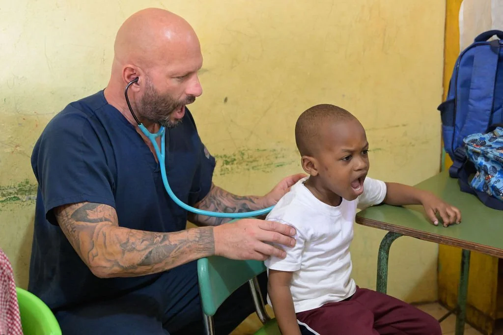 Healthier Futures International volunteer in scrubs listens to a toddler's chest with a stethoscope in 2026