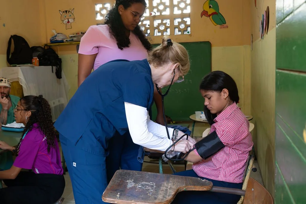 Healthier Futures International volunteer in scrubs takes a girl's blood pressure in a classroom at 2026 clinic