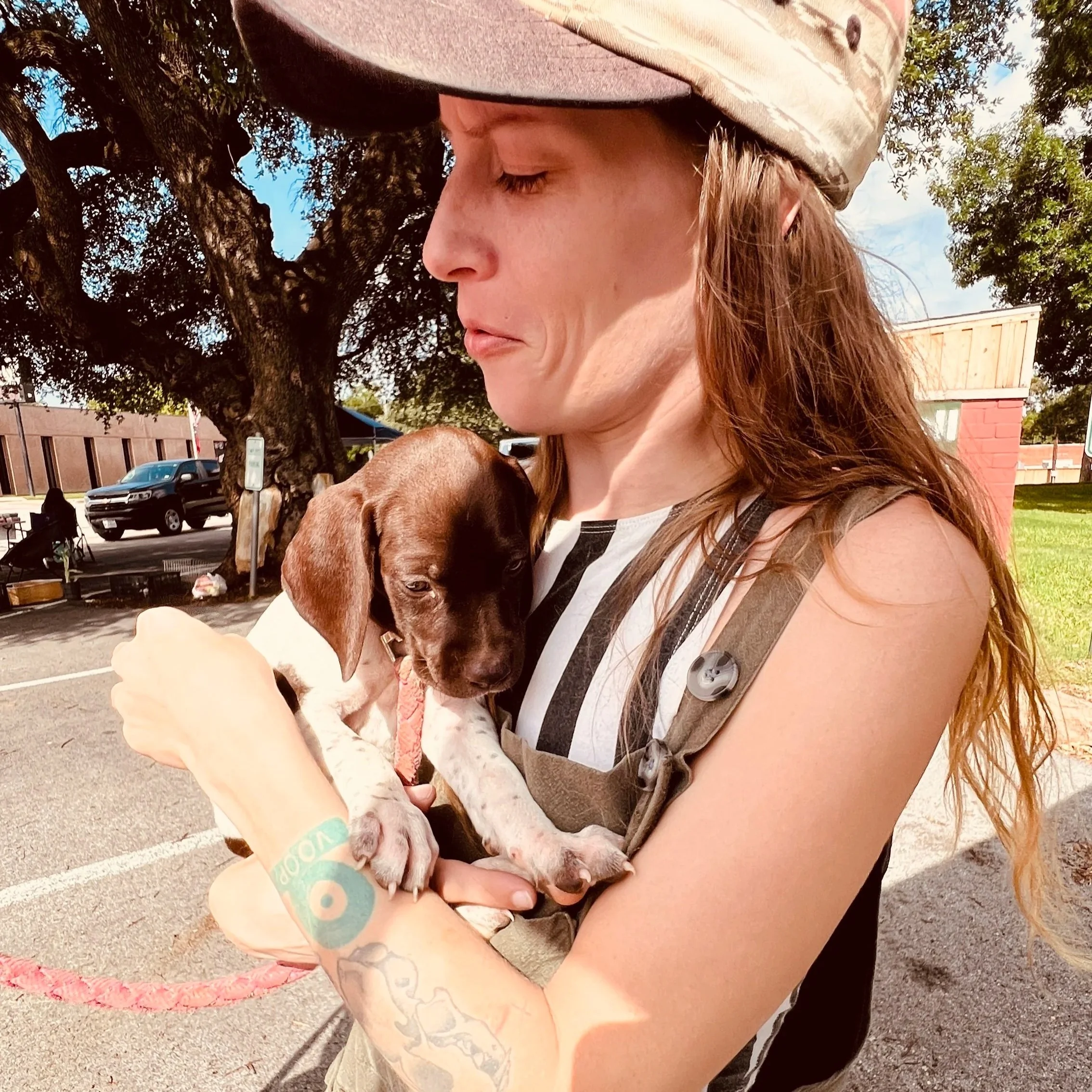A woman with long red hair wearing a camouflage hat, holding a brown and white puppy with a green wristband, outdoors with trees, parked cars, and a building in the background.