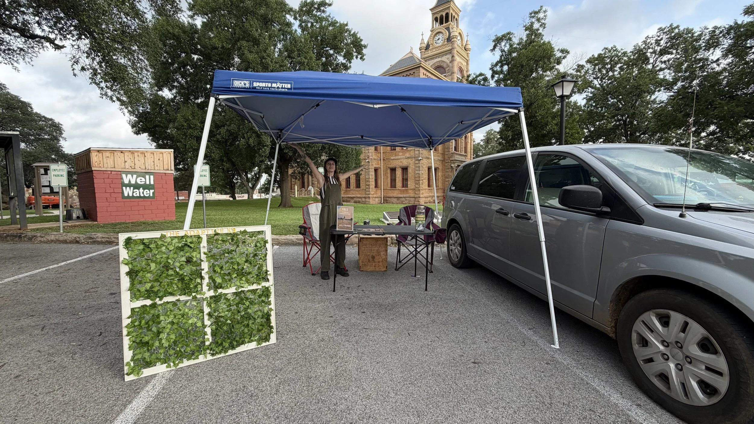 A woman standing under a blue canopy tent at an outdoor market booth with plants and informational signs, next to a gray vehicle, in front of a historic building with a clock tower.