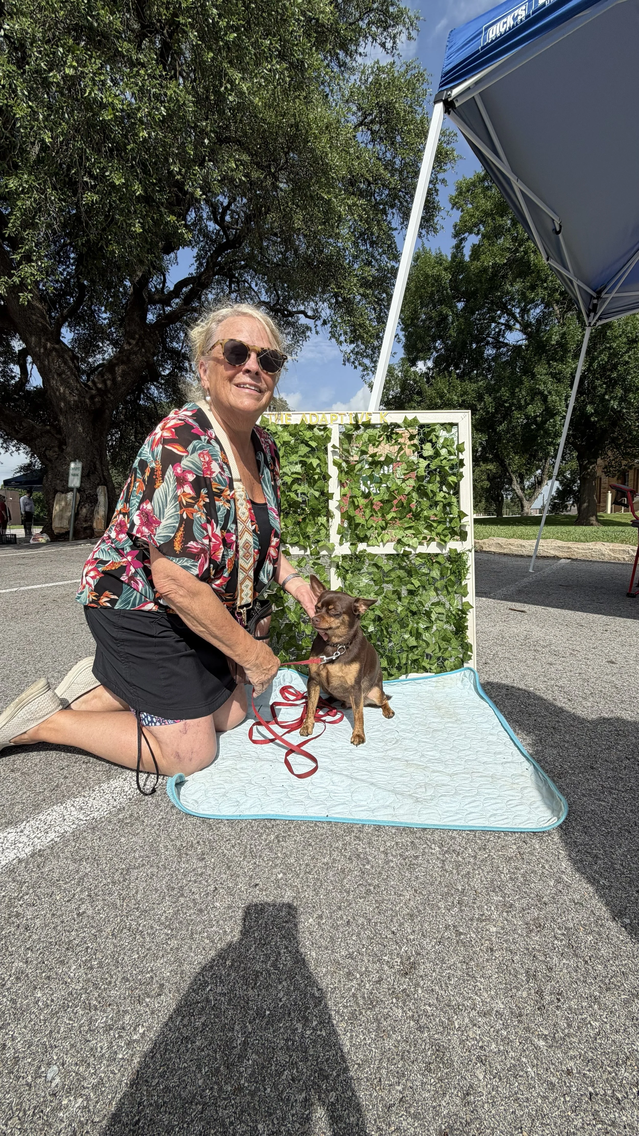 An elderly woman with blonde hair, sunglasses, and a floral shirt kneeling on a mat, smiling with a small brown dog sitting in front of her, outdoors under blue sky, with trees and a display with green plants behind them.