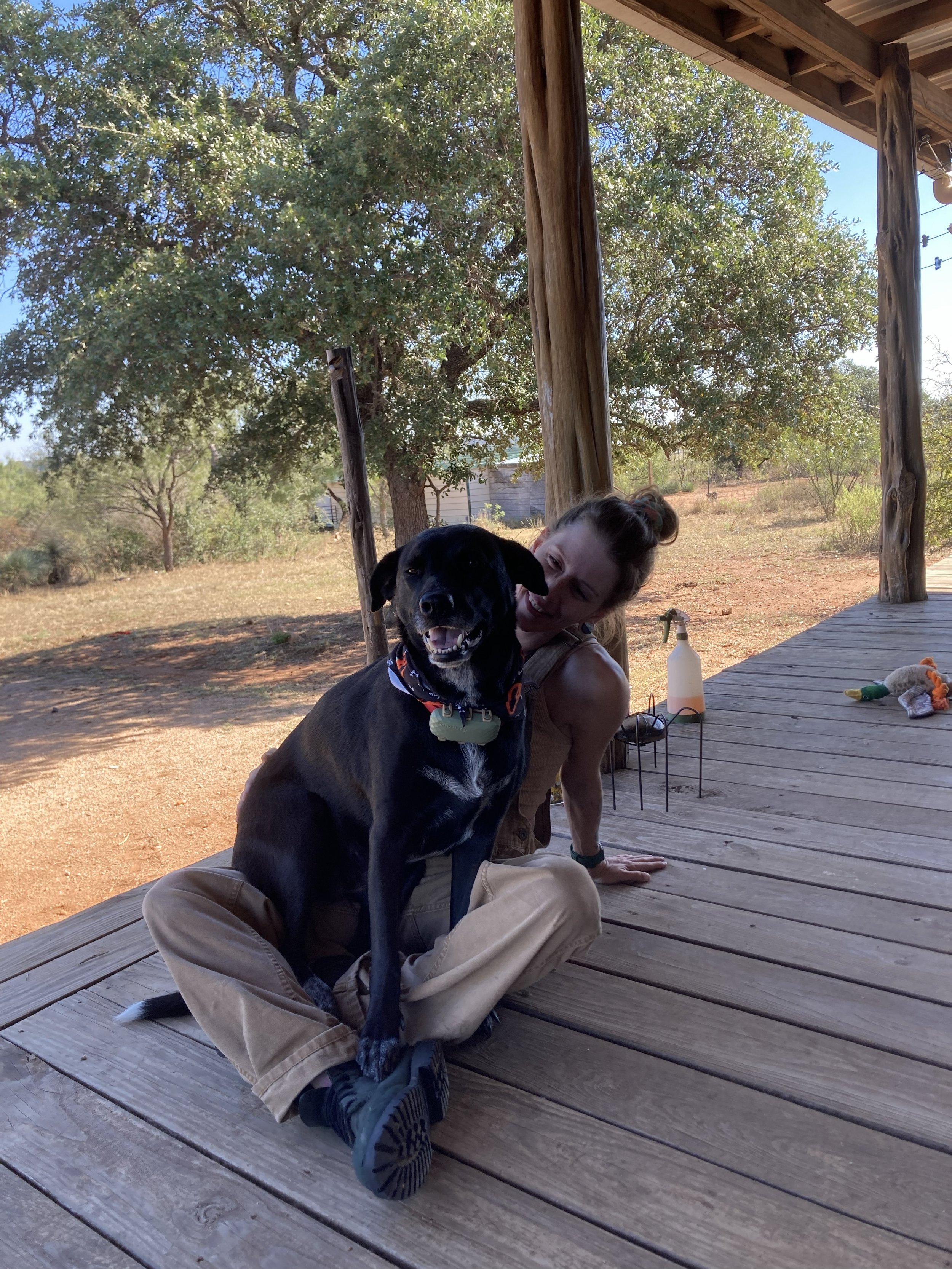 A woman sitting on a wooden porch with a black dog and smiling. There are trees in the background and some toys on the porch.