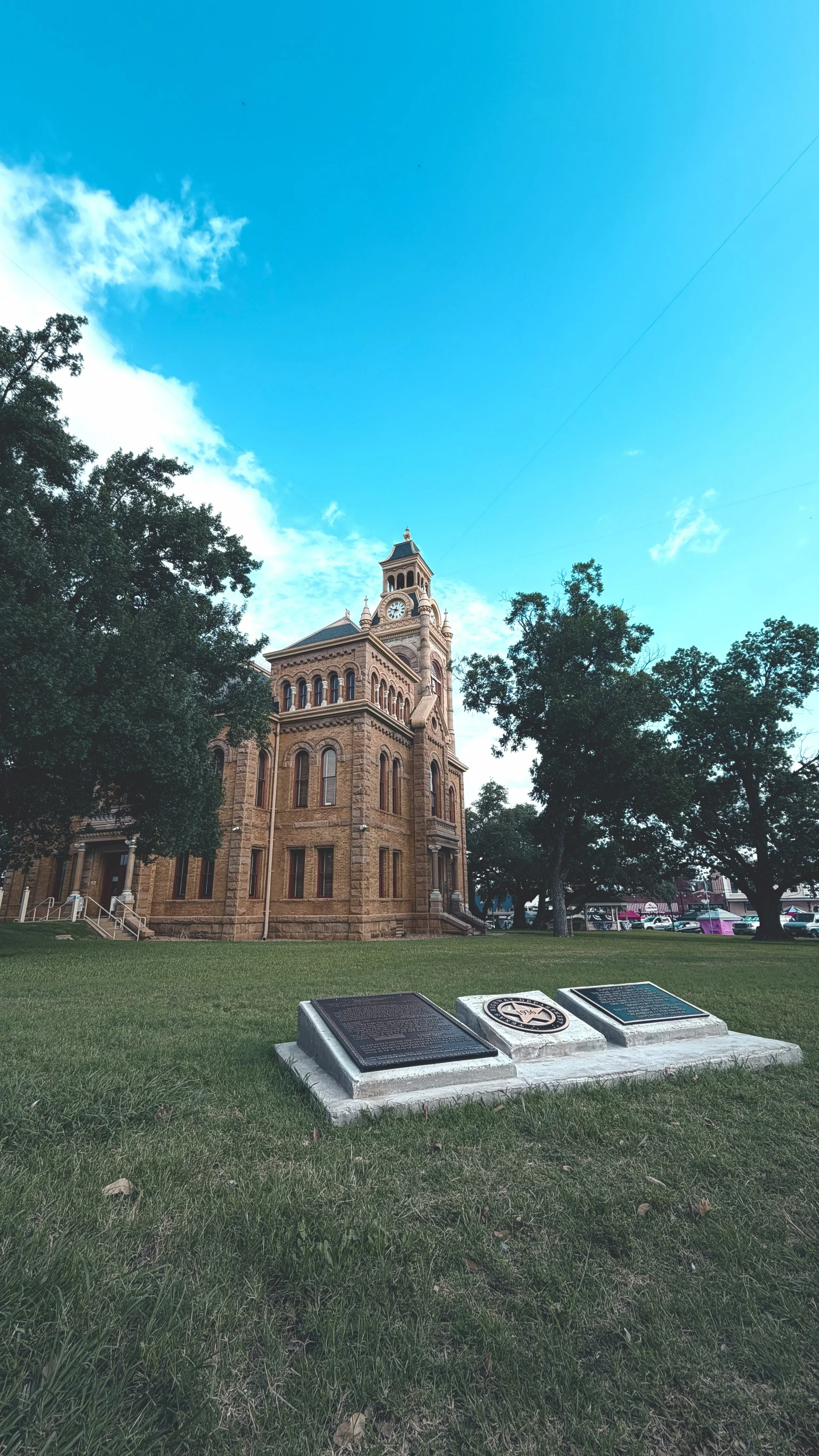 Historic clock tower building with trees and green lawn in the foreground, plaques on a stone marker, and a blue sky with some clouds.