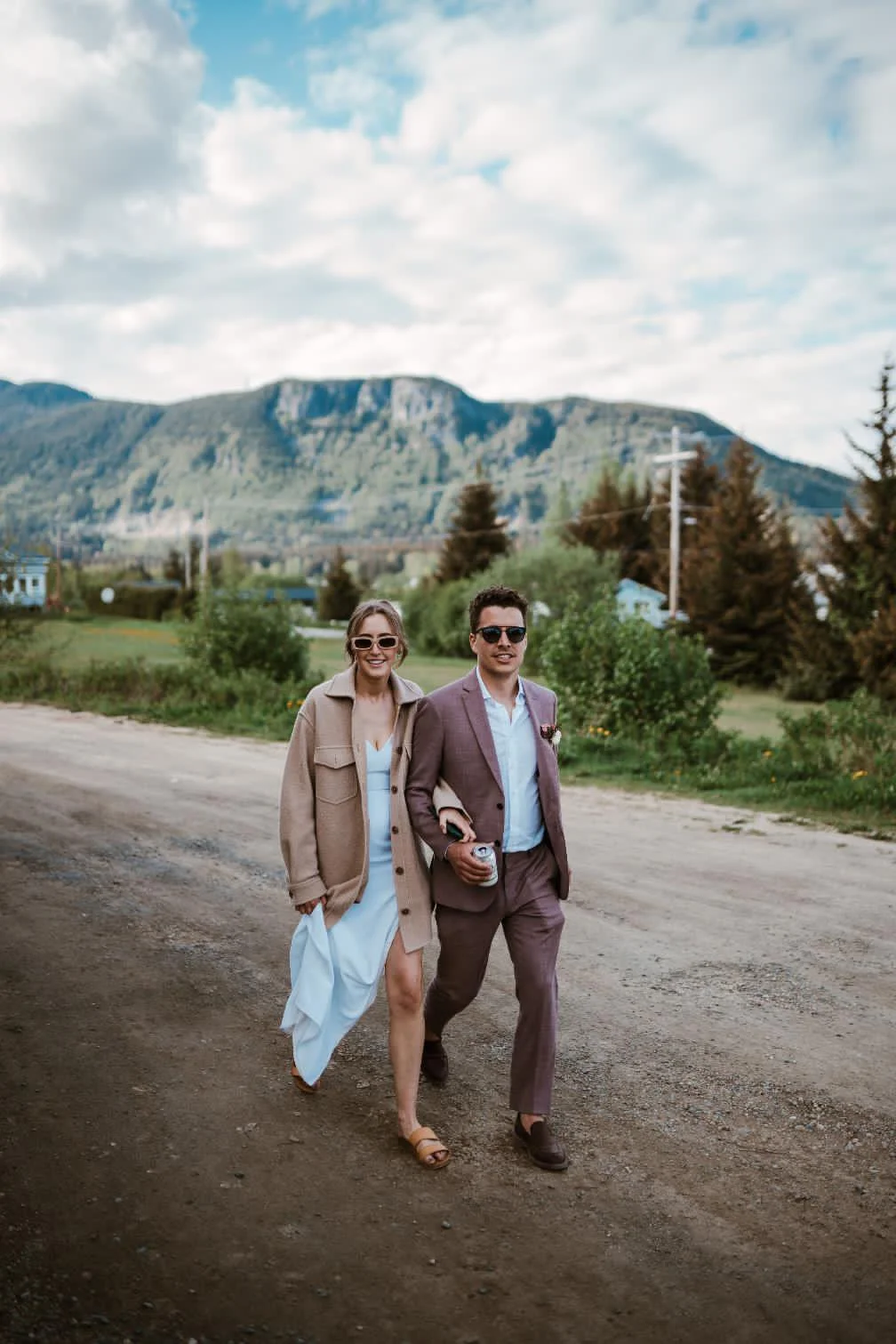 A couple dressed in semi-formal attire walking outdoors on a dirt path, with mountains, trees, and a cloudy sky in the background.