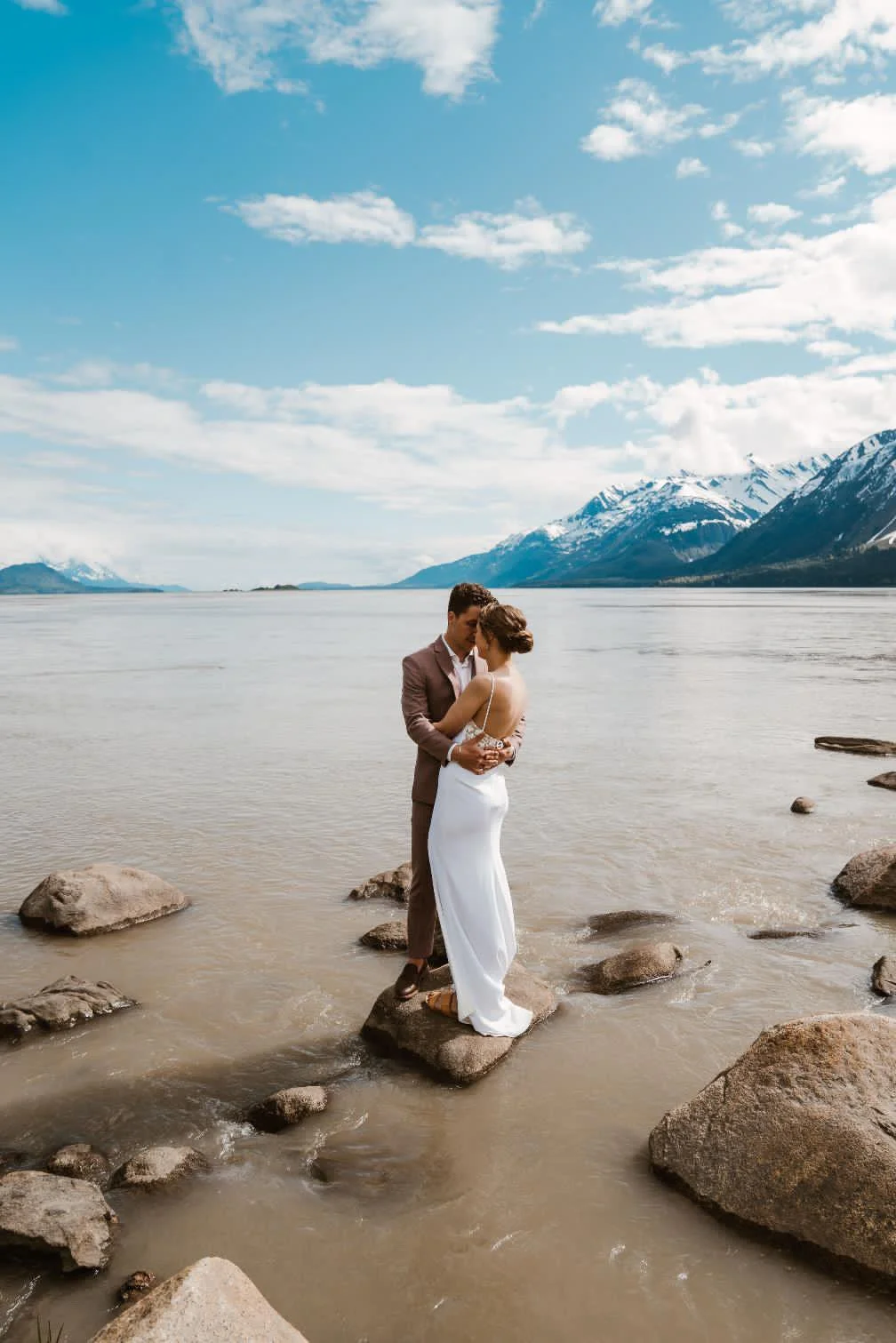 A couple in wedding attire standing on rocks at the edge of a lake with mountains and a partly cloudy sky in the background.
