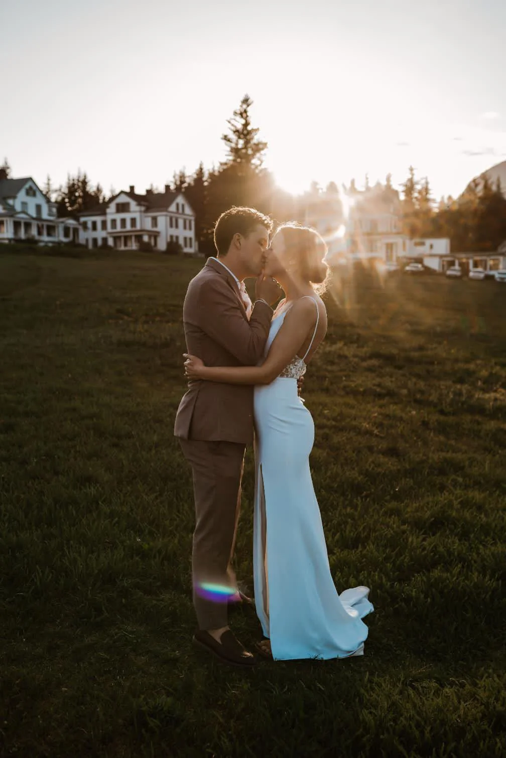 A couple in wedding attire sharing a kiss and embracing outdoors at sunset.
