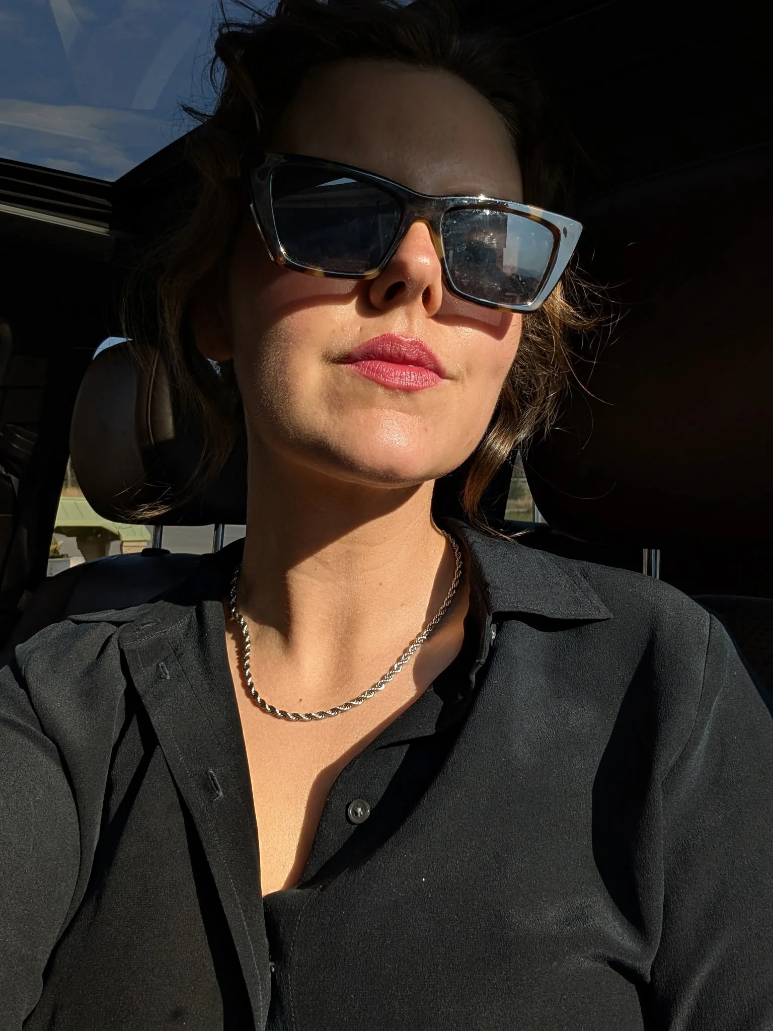 A woman wearing black sunglasses and jewelry, sitting in a vehicle with sunlight on her face with a perfect spray tan.