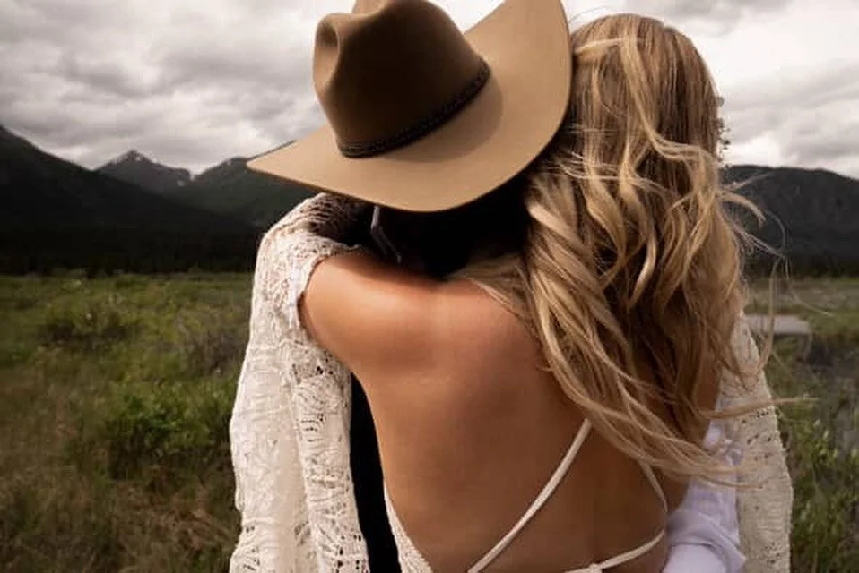 Two women hugging outdoors in a grassy landscape with mountains and cloudy sky in the background, one woman wearing a wide-brimmed hat and lace dress, the other with long wavy hair and white clothing with a perfect spray tan.