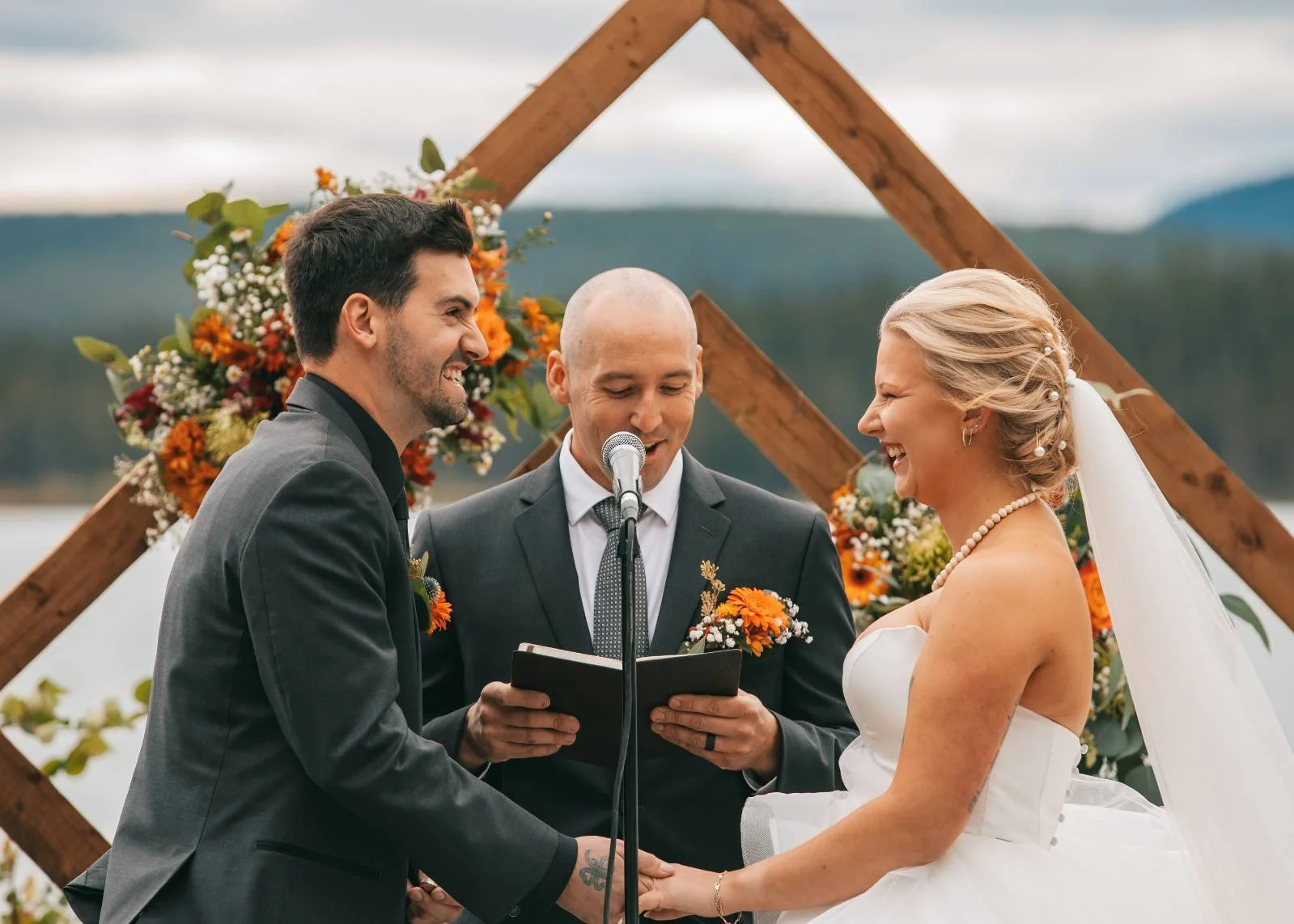 Bride and groom holding hands during their outdoor wedding ceremony with officiant, with a lakeside and forested mountains in the background, decorated with flowers and wood arch with a perfect spray tan.