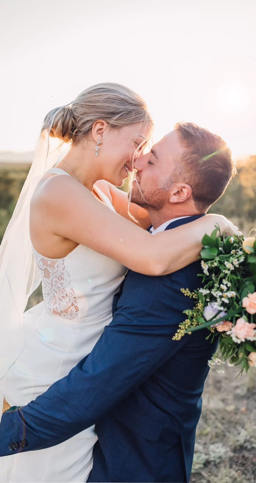A bride and groom embrace and smile during sunset, with the bride wearing a white lace wedding dress and jewelry, and the groom in a dark suit holding a bouquet of flowers with a perfect spray tan.
