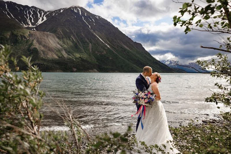 A bride and groom sharing a kiss by a mountain lake, surrounded by greenery.