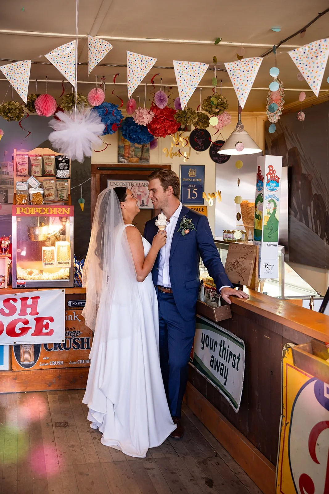 Bride and groom sharing ice cream at a festive ice cream stand decorated with bunting, pom-poms, and paper lanterns, inside a lively venue with a perfect spray tan.