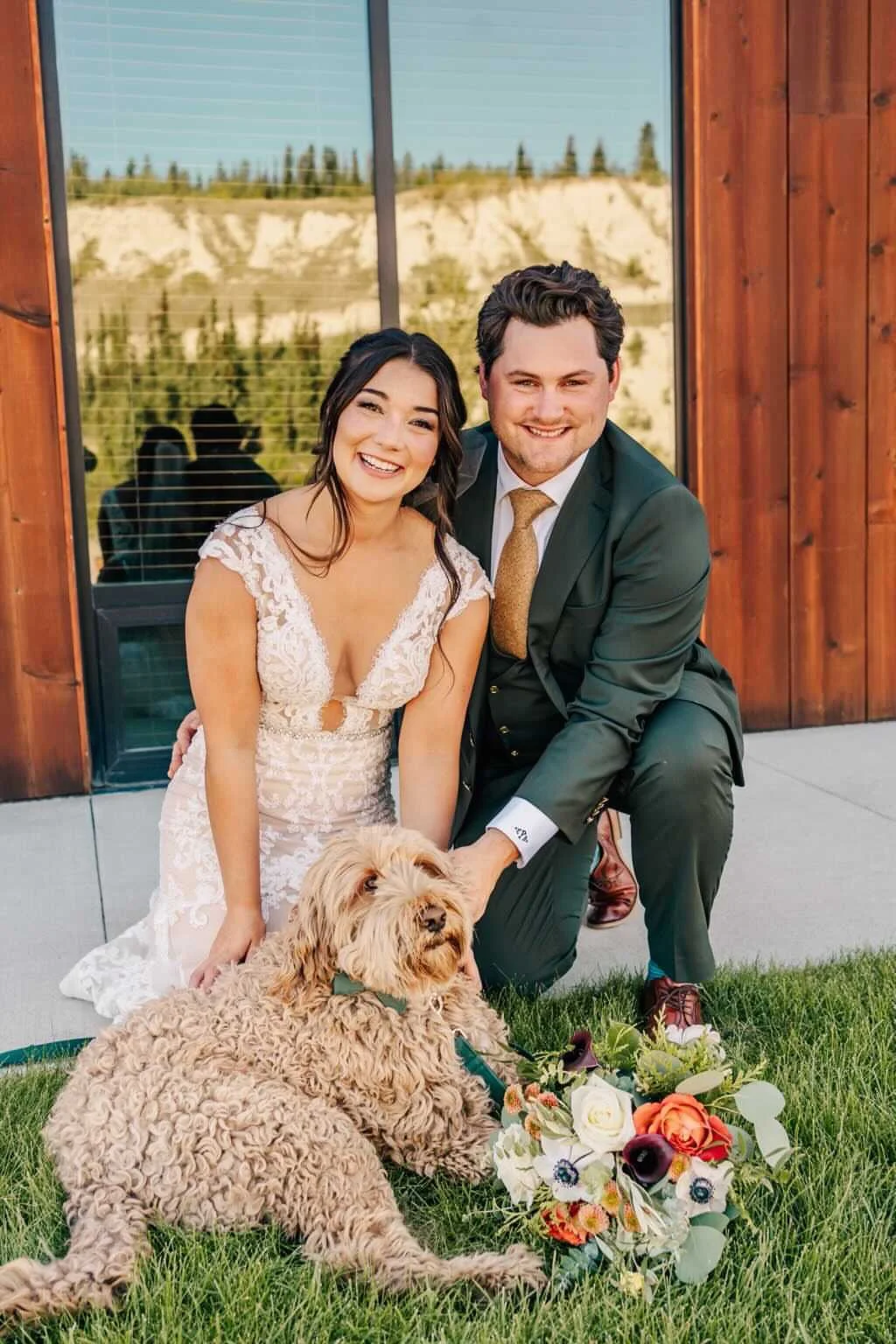 A smiling bride and groom kneel on the grass outside a building with large windows, accompanied by a large, curly-haired dog lying next to a colorful bouquet of flowers.