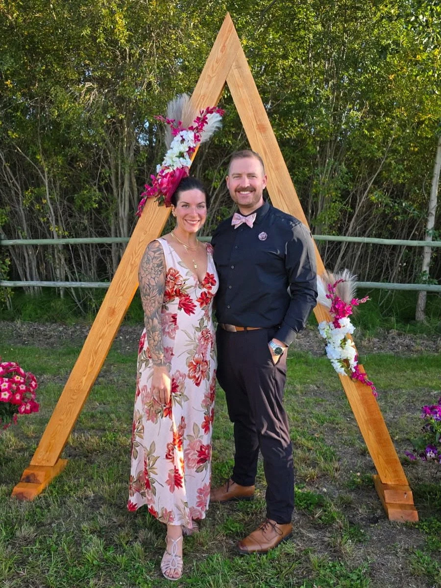 A smiling man and woman standing side by side outdoors in front of a wooden A-frame decorated with pink and white flowers, greenery, and pampas grass, during daytime, dressed in semi-formal attire with a perfect spray tan.