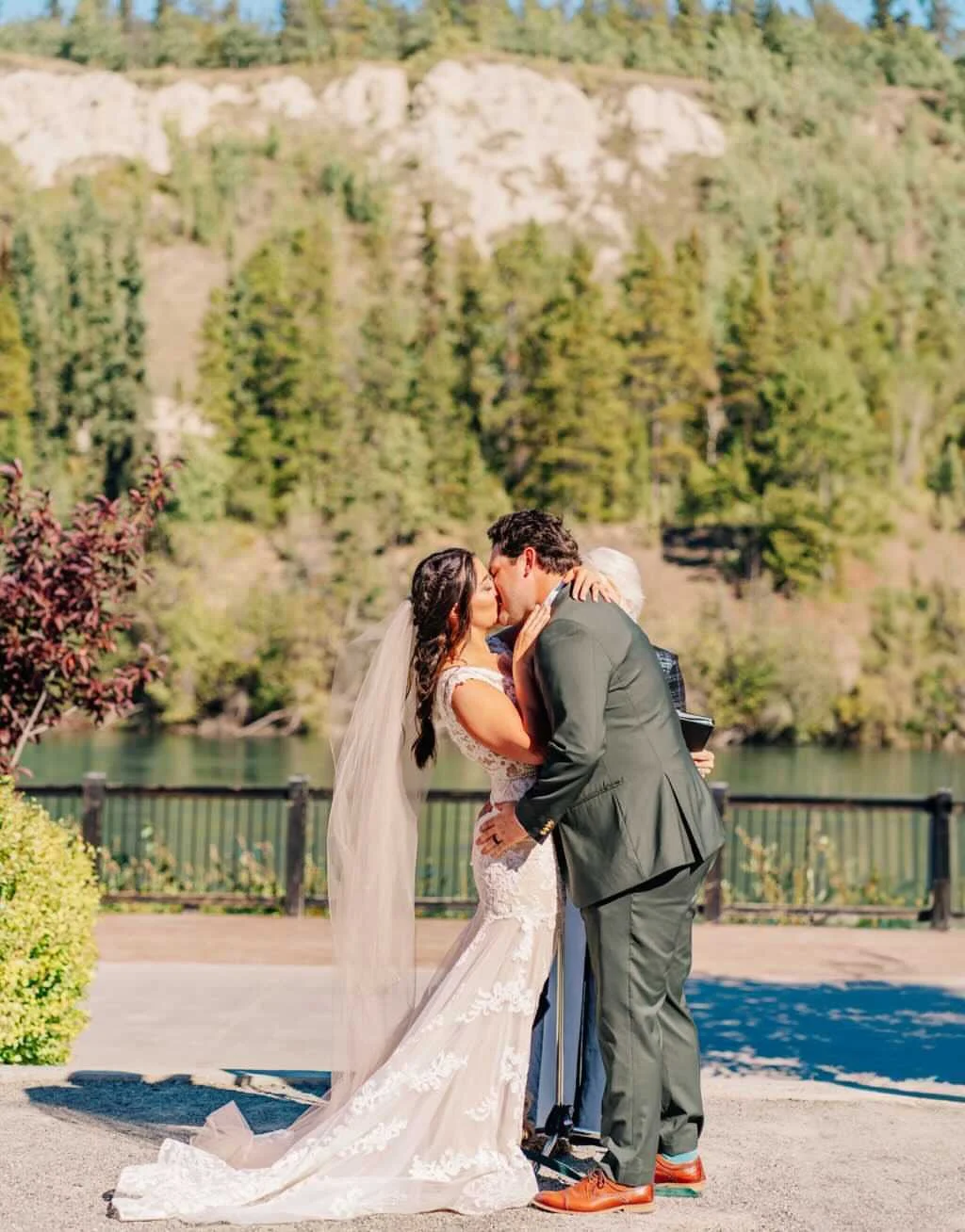 A bride and groom share a kiss outdoors during their wedding ceremony, with a scenic backdrop of trees, a river, and mountains and she has a perfect spray tan.