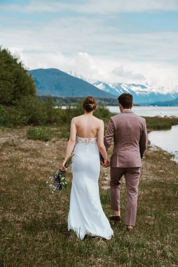 A bride and groom walking hand in hand along a grassy riverside, with mountains and water in the background with a perfect spray tan.