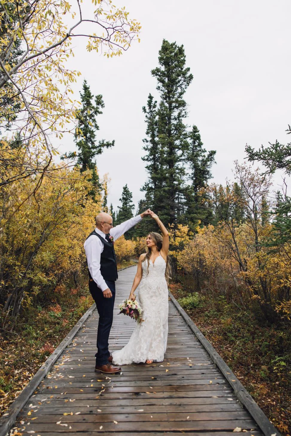 A wedding couple dancing on a wooden pathway surrounded by autumn foliage, with tall pine trees in the background with a perfect spray tan.