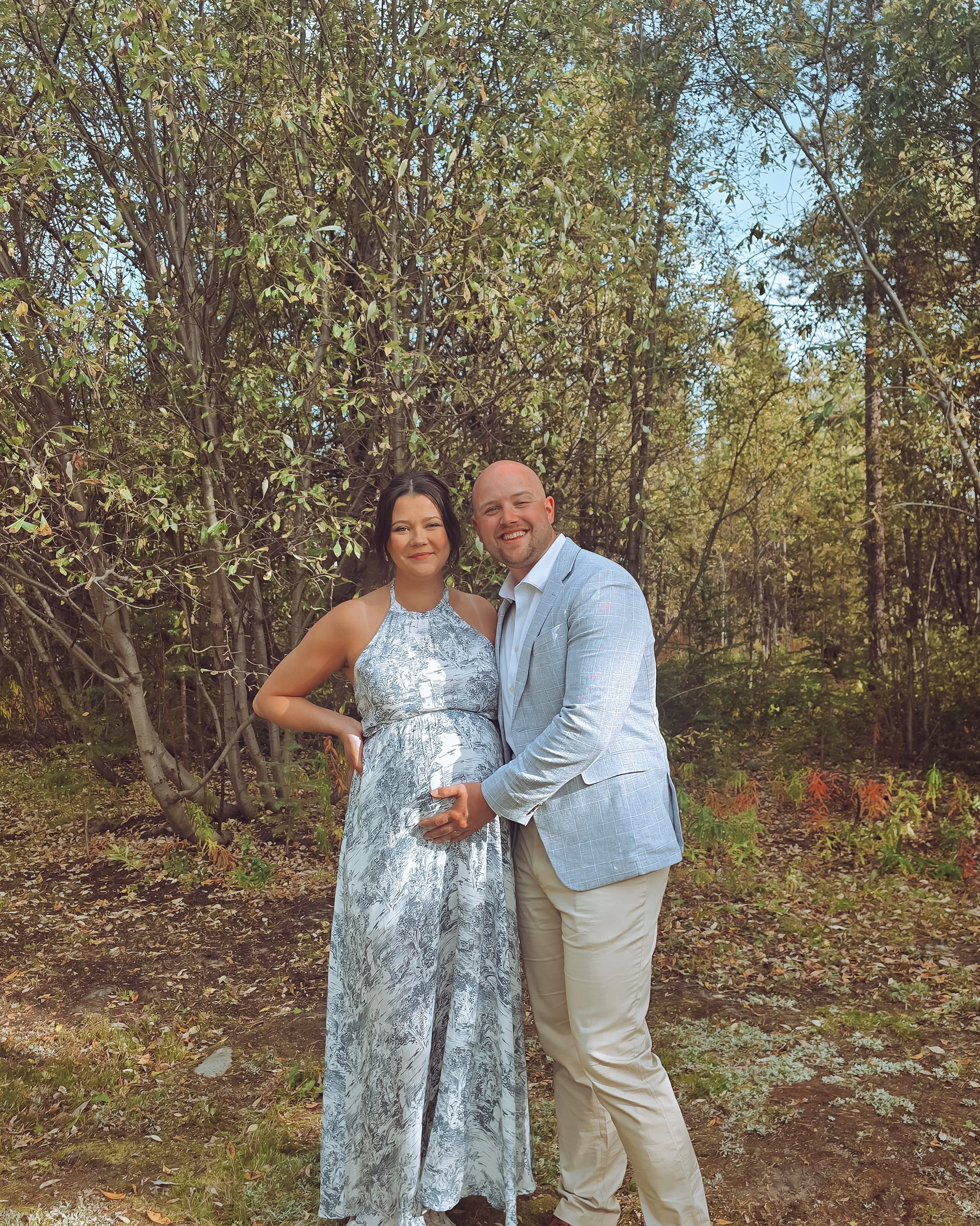 A pregnant woman in a patterned maxi dress standing next to a man in a light gray blazer and beige pants, both smiling and holding her baby bump outdoors with trees in the background with a perfect spray tan.