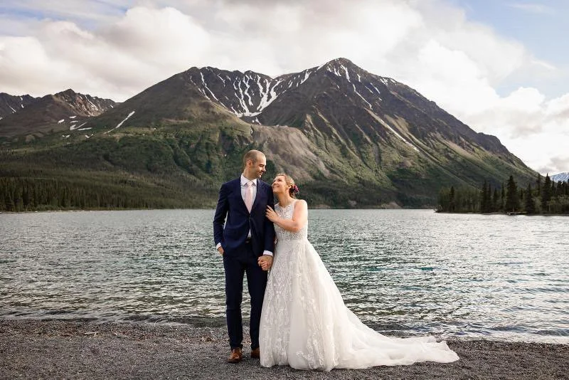 Bride and groom holding hands by a lake with mountains in the background