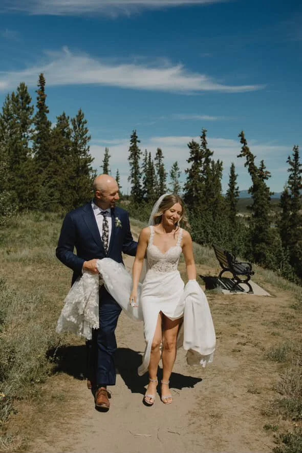A bride and groom walk outdoors on a sunny day, with tall trees and a blue sky in the background. The bride is wearing a white wedding dress, and the groom is in a dark suit. They are smiling and holding their shoes and dresses while walking on a dirt path.