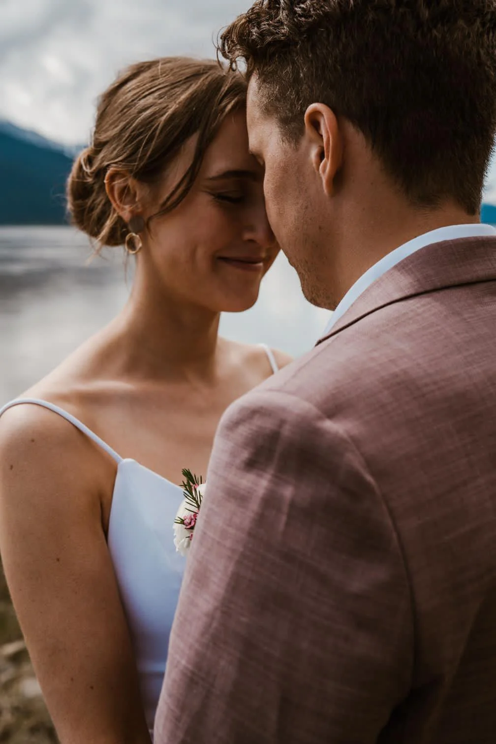 A woman and a man standing close with their foreheads touching, outdoors near a lake with mountains in the background, both smiling softly. The woman is wearing a spaghetti strap dress with a small floral accessory on her chest. The man is dressed in