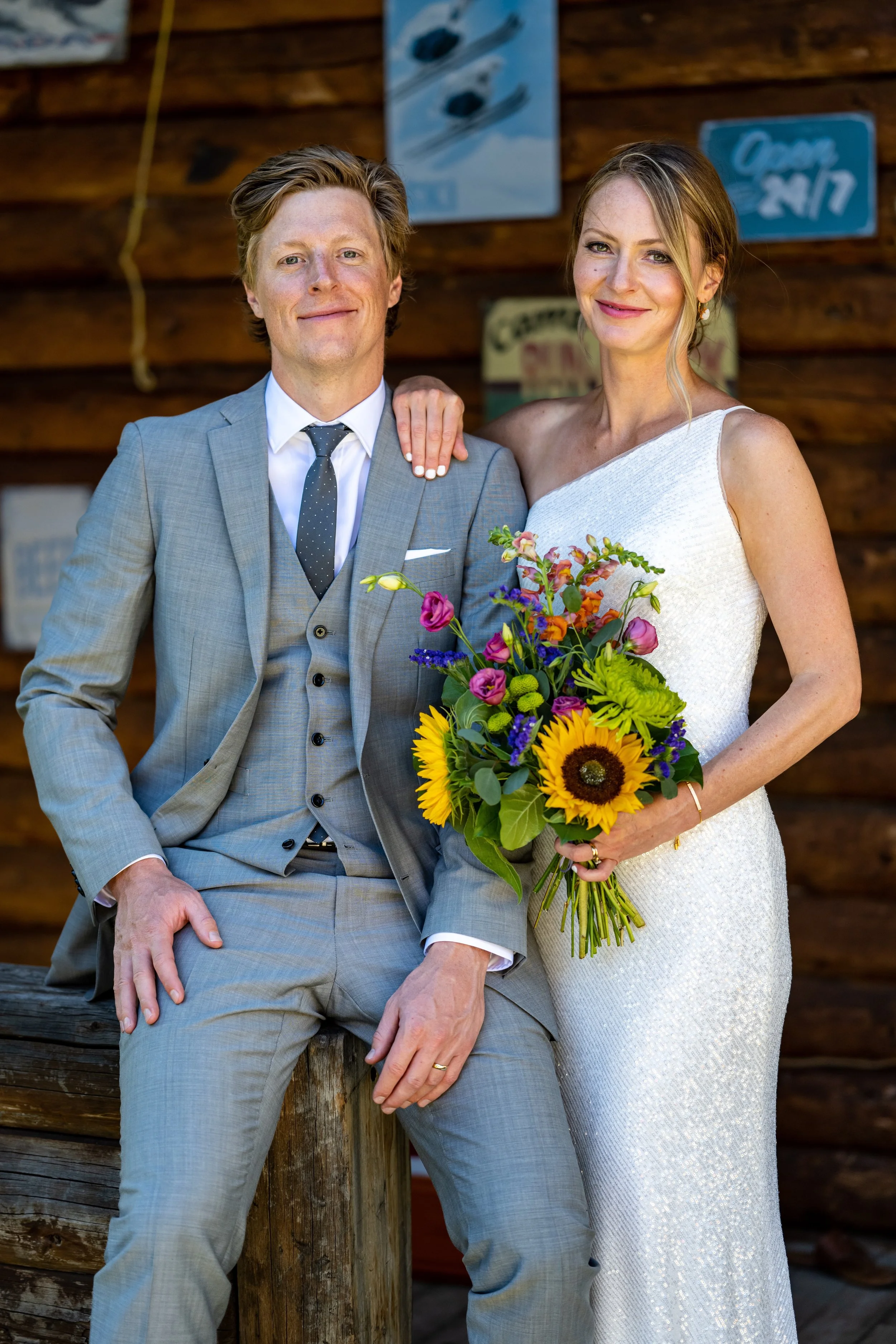 A man in a gray suit and a woman in a white dress, holding a bouquet of sunflowers and colorful flowers, posing outdoors against a wooden wall with a perfect spray tan.