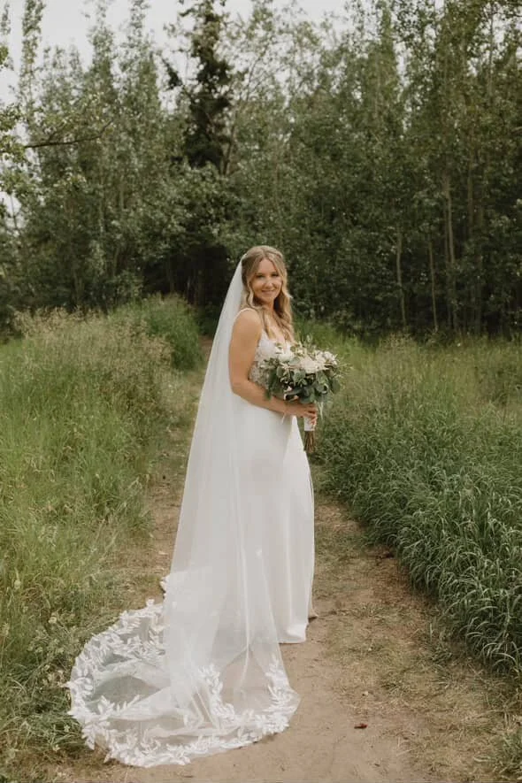 A smiling bride in a white wedding dress with a long veil holding a bouquet of flowers on a dirt path surrounded by greenery and trees.