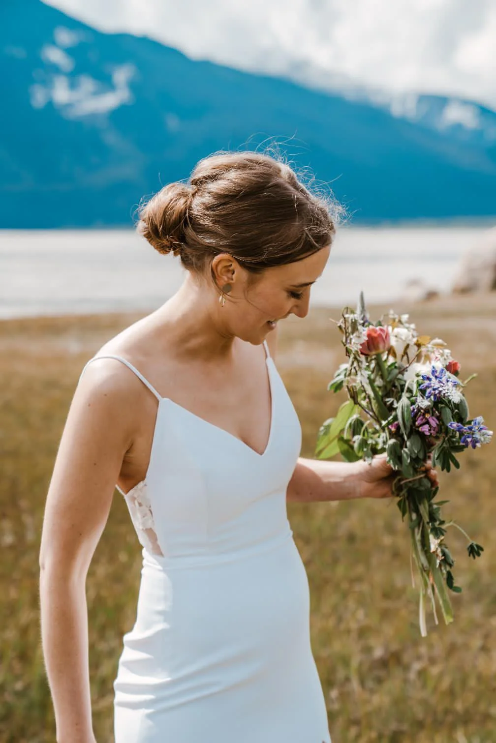 A woman in a wedding dress holding a bouquet of flowers outdoors with mountains and water in the background.