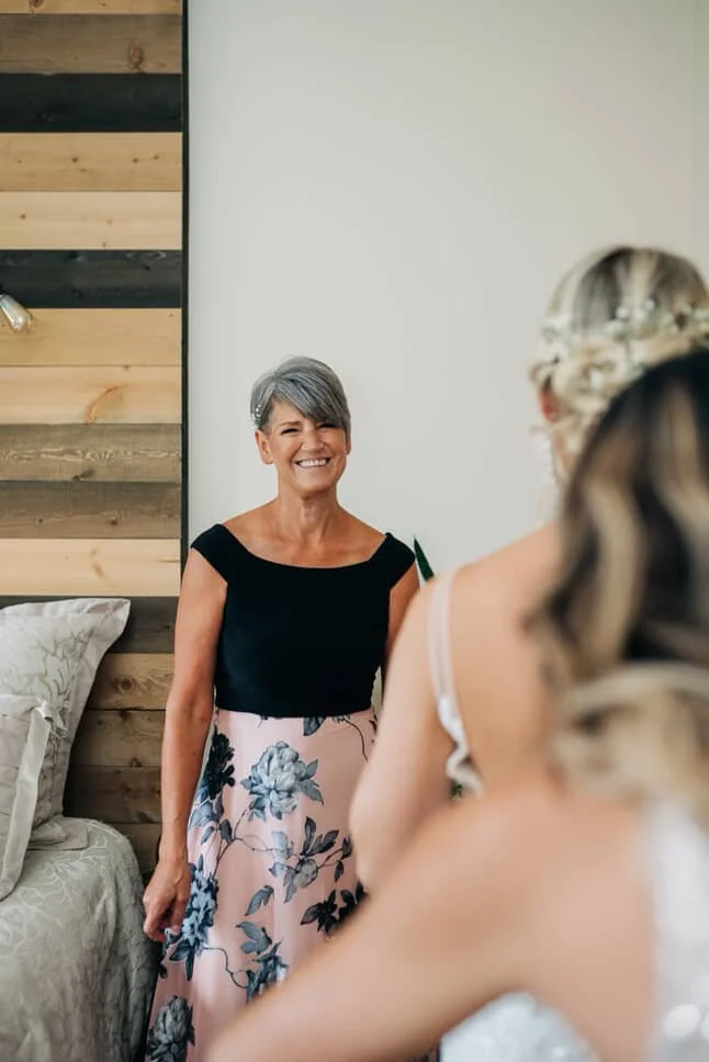 A woman with short gray hair, wearing a black top and a pink floral skirt, smiling as she looks at a bride with her own glowing tan.