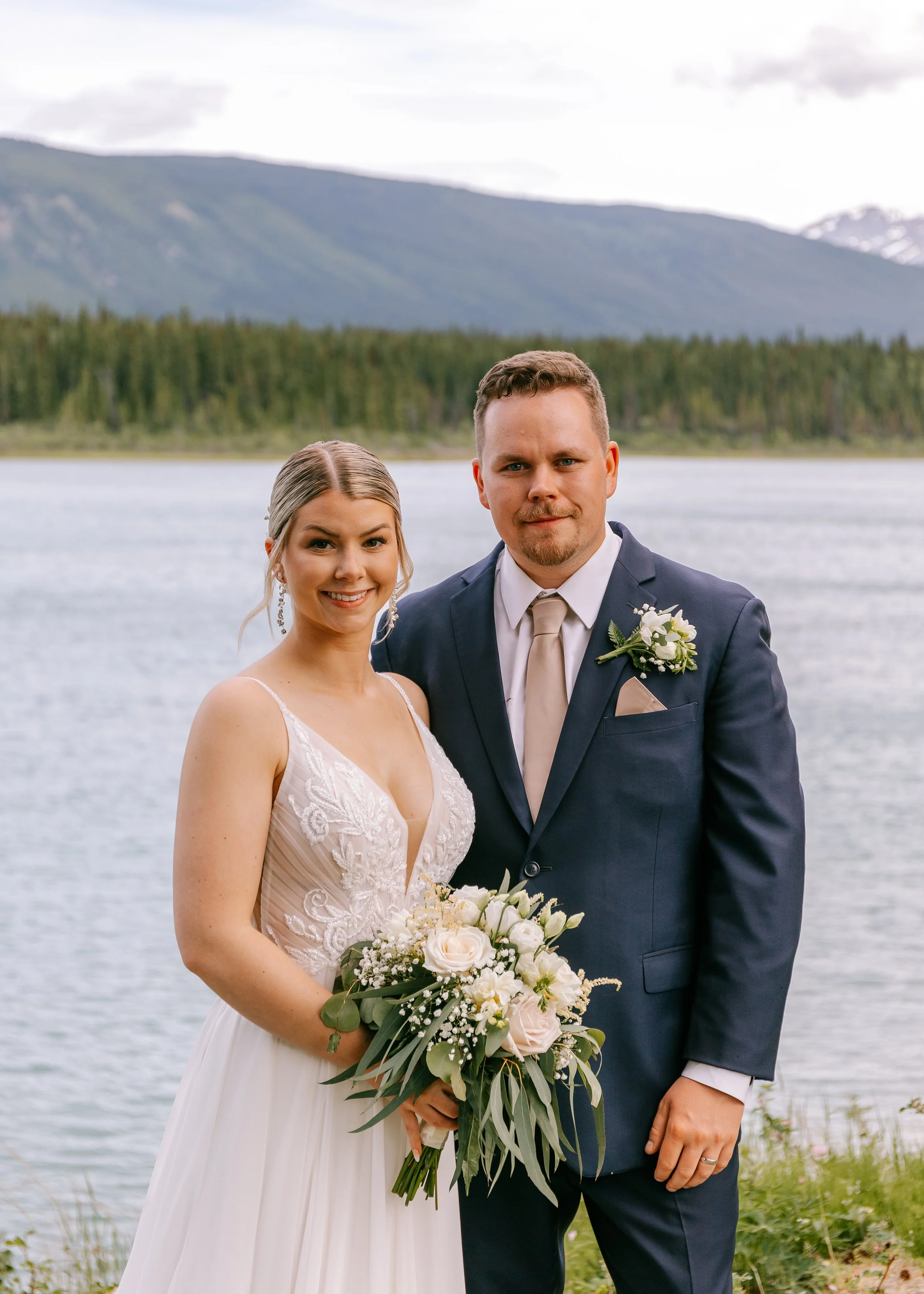 Bride and groom standing outdoors near a lake, with mountains and trees in the background, during their wedding celebration with a perfect spray tan.