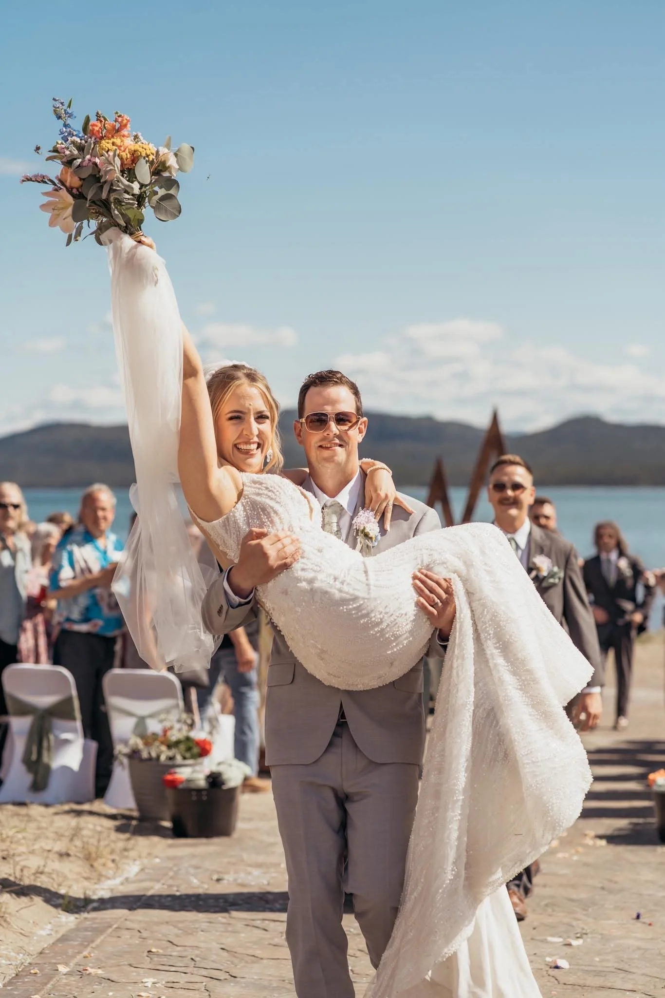 A groom carrying a smiling bride in his arms outdoors during a wedding ceremony, with friends and family in the background, near a body of water with mountains in the distance with a perfect spray tan.