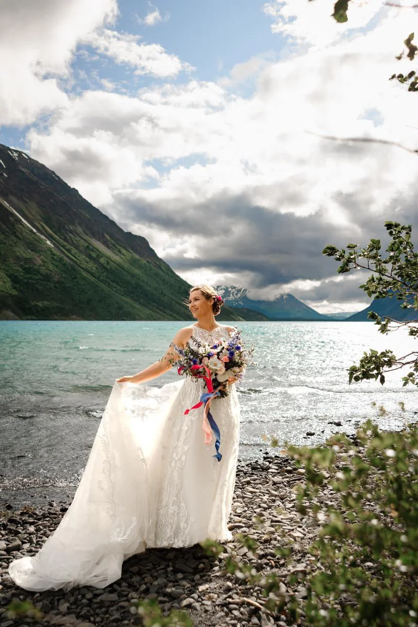 Bride in a white wedding dress holding a colorful bouquet, standing on a rocky lakeshore with mountains and cloudy sky in the background.