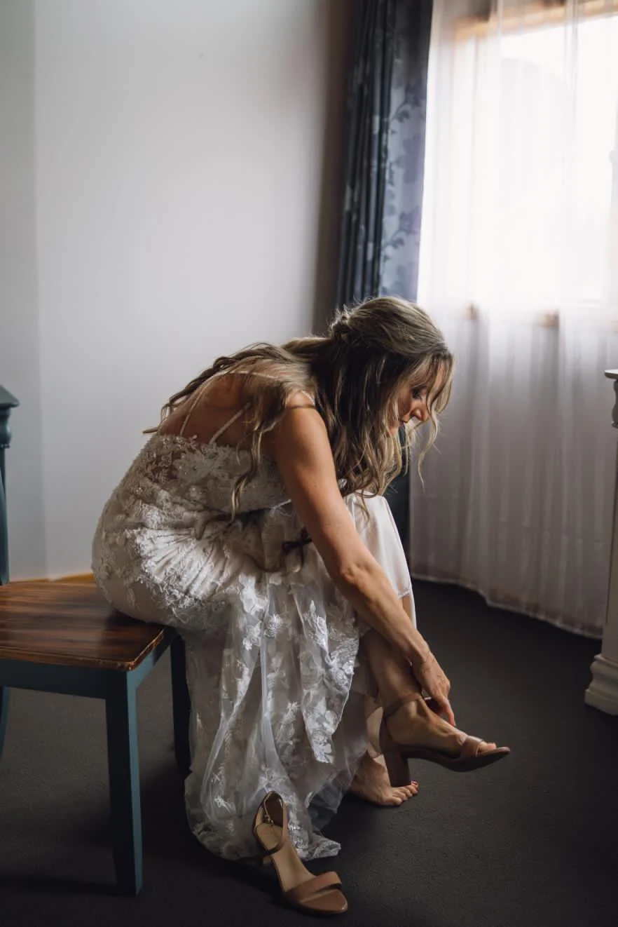 A woman in a wedding dress sits on a chair, putting on her shoe in a softly lit room with a window and curtains with a perfect spray tan.
