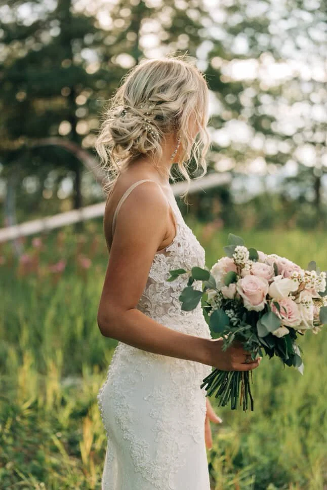 A bride standing outdoors holding a bouquet of pink and white roses and greenery, with trees and blurred background with a perfect spray tan.
