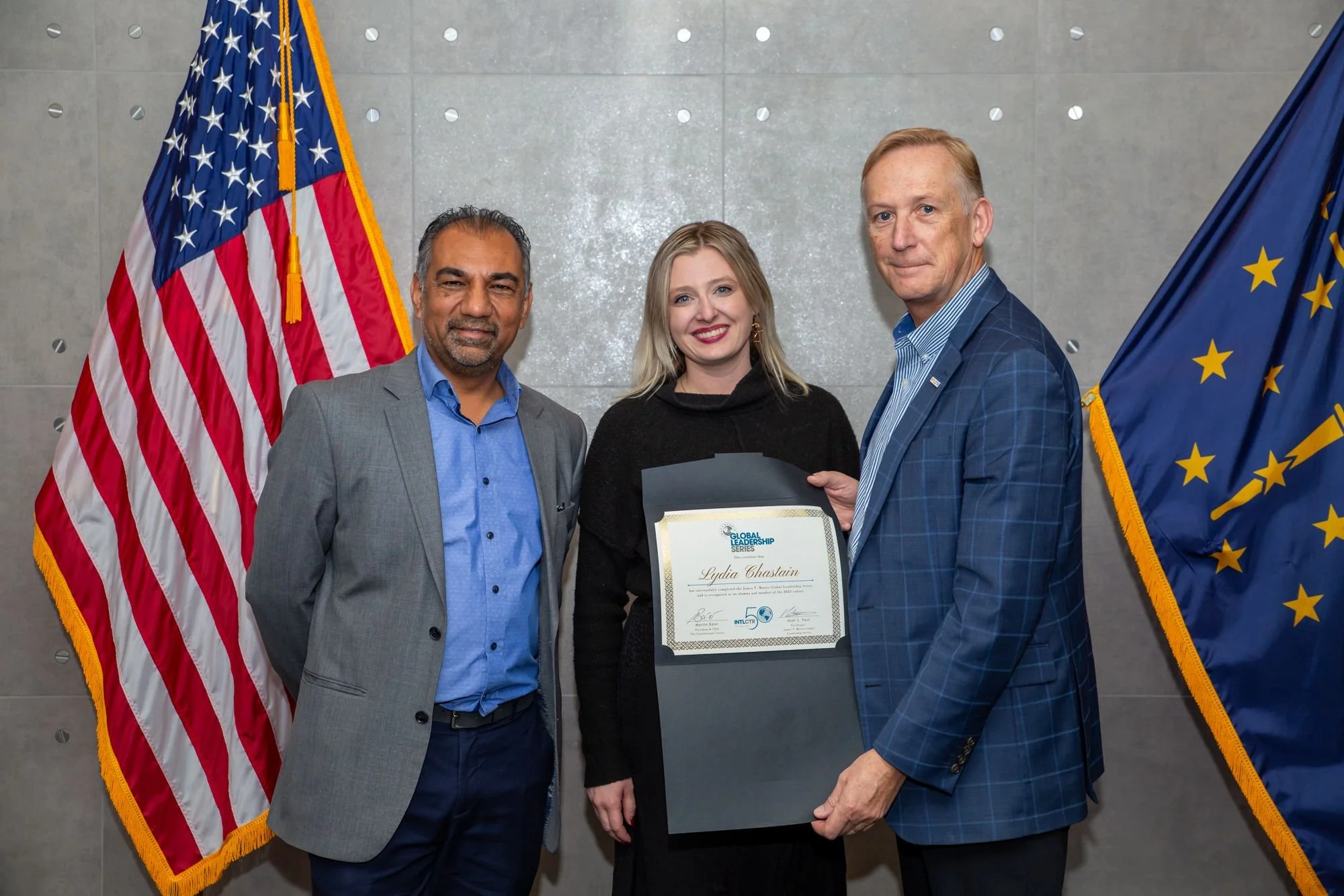 Three people standing together, one woman in the middle holding a certificate, with two men on either side, all dressed in business attire, USA flag and Indiana flag in the background.